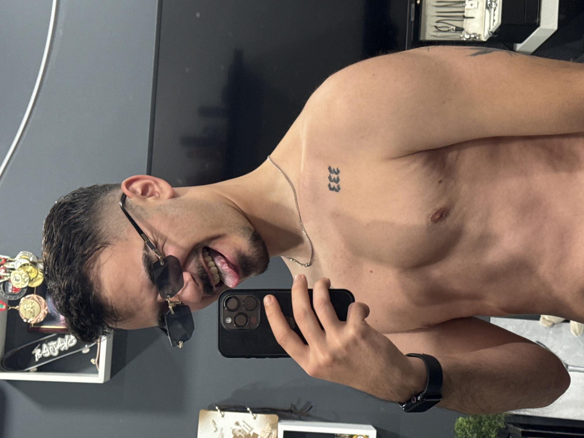 A shirtless man with a dark beard and sunglasses takes a selfie with a black iPhone in what appears to be an indoor setting, possibly a bathroom. The man is looking directly at the camera with his tongue slightly sticking out. He has a tattoo of the number "333" on his right clavicle and a silver chain around his neck. A black smartwatch is visible on his left wrist. Behind the man, there is a grey wall and a shelf containing various objects, including a white-framed display with coins and other items. There is no information available to determine the specific location in Sofia, Bulgaria, as no identifying landmarks are present in the image.