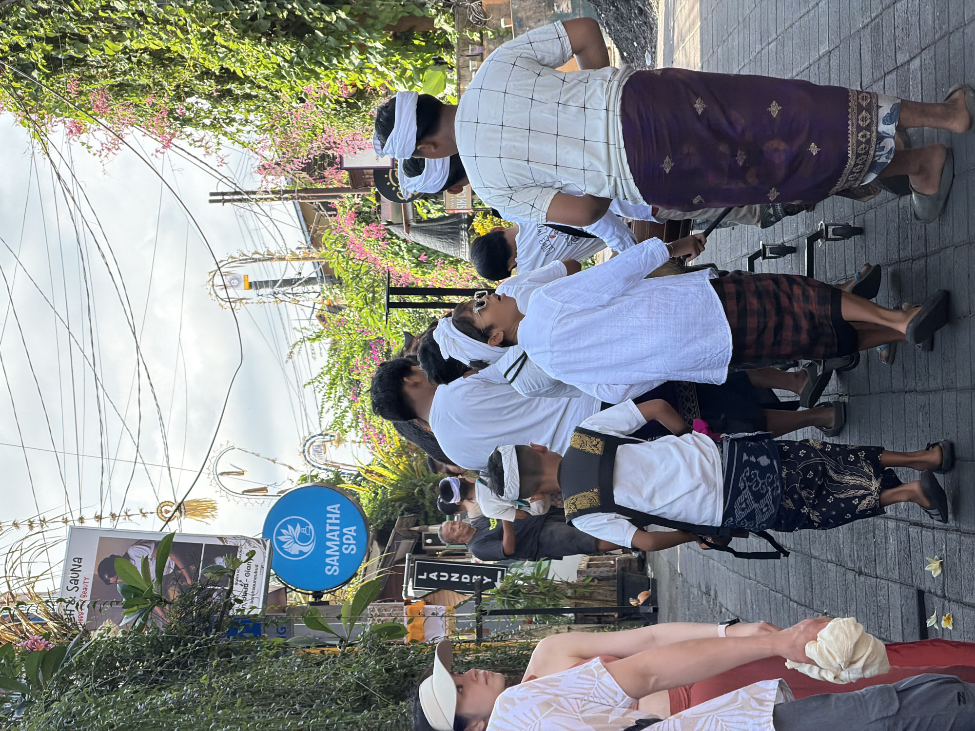 In Ubud District, Indonesia, a group of people, including several young boys dressed in white tops and traditional sarongs, walk down a paved street lined with lush greenery and flowering vines. One boy in the foreground wears a white checked shirt and a dark purple sarong with gold embroidery. To his left, another boy in a white shirt and sunglasses looks off to the side. A woman in a white visor and floral top is visible on the left, and other individuals are seen in the background. Overhead, a tangle of electrical wires crisscrosses the sky. A circular blue sign with the text "SAMATHA SPA" and a woman's silhouette is mounted on a pole. Another sign advertising "LAUNDRY" is also visible. Some of the individuals are pushing a wheeled cart. The overall scene suggests a busy street in a tropical urban environment, possibly during a cultural event or procession.