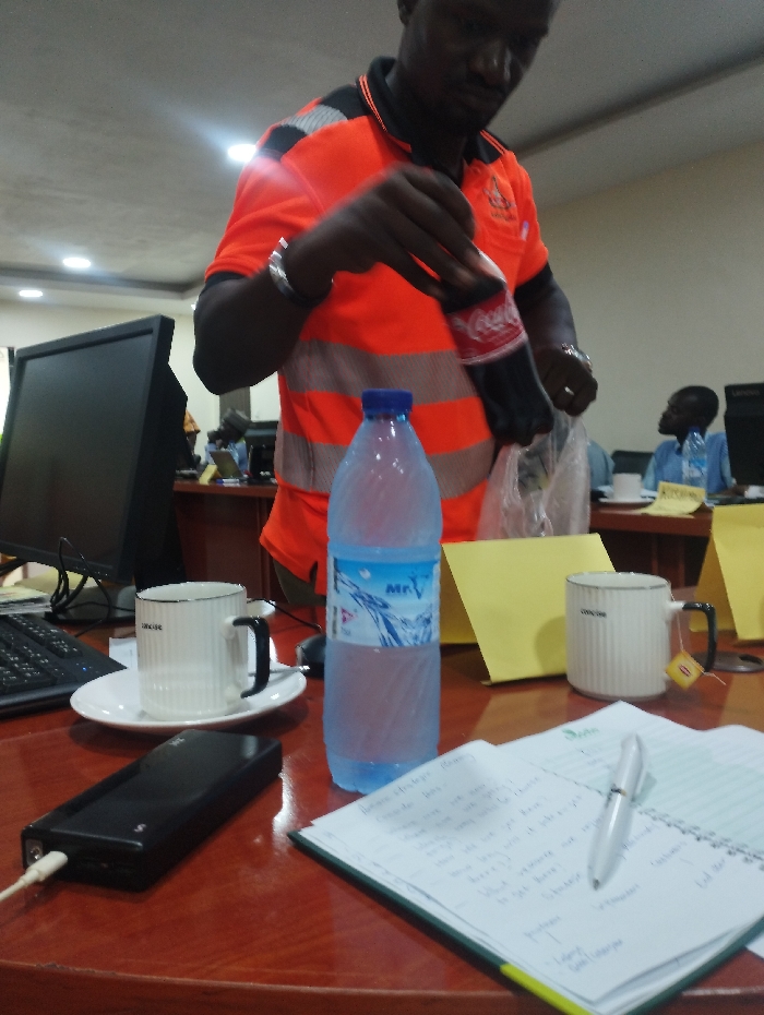 The image depicts a scene inside a room in Ashaka, Nigeria (latitude: 10.929, longitude: 11.476), likely an office or meeting space. A man wearing an orange safety vest, holds a glass bottle of Coca-Cola in his right hand while pouring the beverage into a clear plastic bag in his left hand. He is standing at a wooden table that holds several objects, including a water bottle, two coffee mugs, a black power bank, and a notebook with a pen. There is also a computer keyboard and a desktop monitor visible in the background. A second person, also wearing a safety vest, is sitting at the table to the right, outside the frame of the image. The lighting is bright and the room appears well-maintained.
