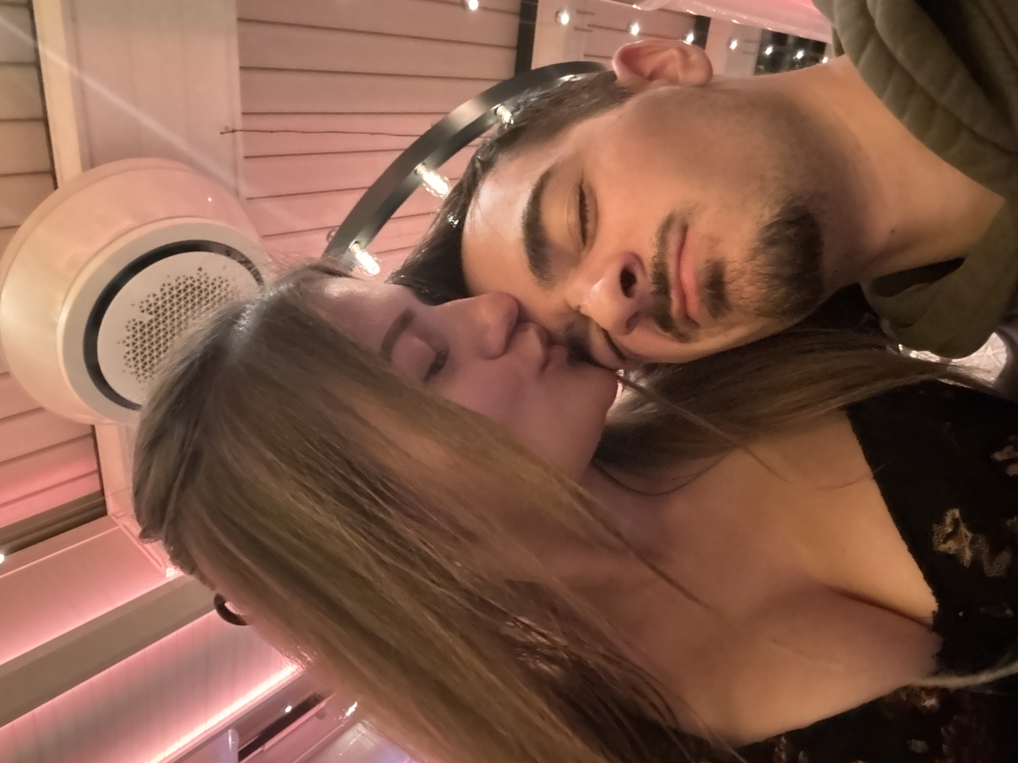 The image captures a close-up, chest-up view of two individuals, a female and a male, engaged in an affectionate interaction within an indoor commercial establishment in Sofia, Bulgaria.
The female, positioned on the left, has long, light brown hair. She wears a dark, low-cut garment, revealing cleavage, on which a faint vertical mark is visible. She leans in, eyes closed, to kiss the male's forehead/temple. The male, positioned on the right, has short dark hair, a mustache, and a goatee. He is wearing a dark green hooded sweatshirt, and his eyes are partially closed.
The background consists of a ceiling constructed from slatted wooden panels, illuminated by prominent pink-hued ambient lighting. A white circular cassette-style air conditioning unit with a central perforated vent is mounted on the ceiling in the upper right quadrant. Behind and above the male's head, a dark, arched decorative fixture embedded with multiple small, clear bulbs is visible. The overall environment suggests a modern restaurant or bar setting during evening hours.