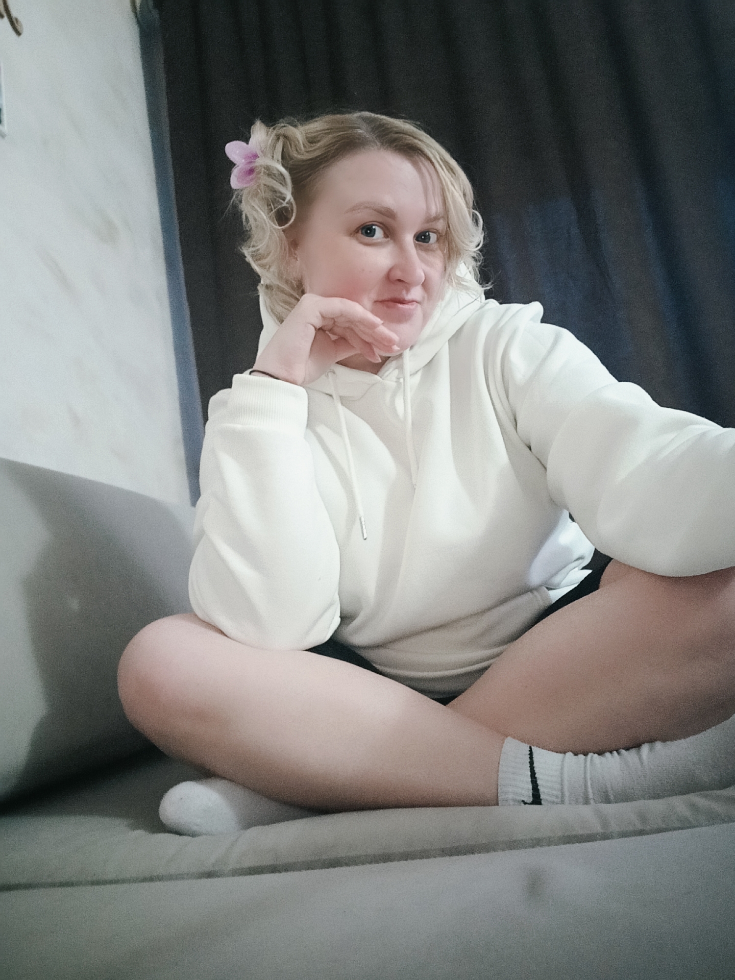 A woman sits comfortably on a light grey sofa in an indoor setting. She has blonde, curly hair, with a small pink flower clip adorning the hair on the left side of her head. Her blue eyes are directed towards the camera, and she has a calm, slightly smiling expression, with one hand resting under her chin. She is wearing a white hooded sweatshirt, dark shorts, and white athletic socks, one of which prominently displays a black Nike swoosh logo. Her legs are crossed in a relaxed pose. In the background, a light-colored wall is visible to the left, and dark curtains are seen to the right, suggesting a cozy room. The lighting is soft and ambient, characteristic of indoor photography. The activity taking place is a posed portrait or selfie. This scene is located in Chaikovsky, Russia.