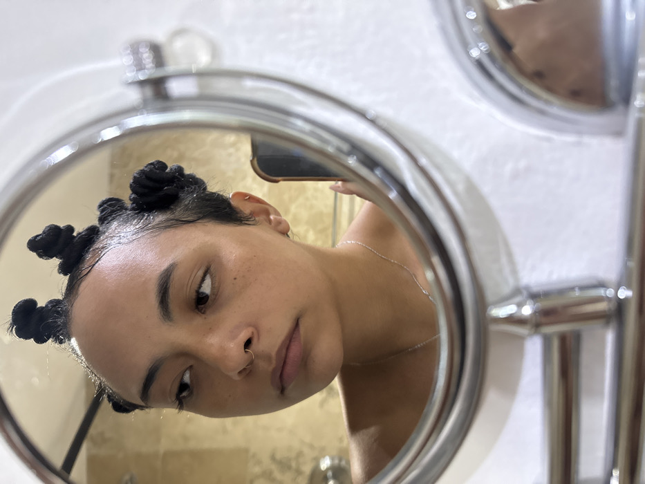 A young individual, appearing female, is reflected in a round, chrome-framed vanity mirror, capturing a selfie. The individual has light brown skin, visible freckles across the nose and cheeks, and dark hair styled into five distinct, small, coiled knot-buns on top of the head, with the hair appearing damp or wet. A thin gold-colored nose ring is present in the right nostril, and a small gold-colored hoop piercing is visible in the reflected left ear. A thin silver-colored chain necklace is worn around the neck. The individual's gaze is directed to the viewer's left, slightly past their own reflection.
The reflection reveals a bathroom setting. A light-colored, possibly beige, tiled wall is visible on the left side of the reflection, while a plain white wall is seen on the right. A chrome towel bar or similar fixture is reflected in the lower background. To the right of the individual's head, a dark-colored smartphone, held by a hand with light-colored fingernail polish, is visible in the reflection, indicating the method of capturing the image. The lower part of the image shows the chrome elements of the mirror's stand or articulation.
No specific visual cues are present within the image to identify a particular location in the city of Taxco, Mexico; the interior setting is generic.