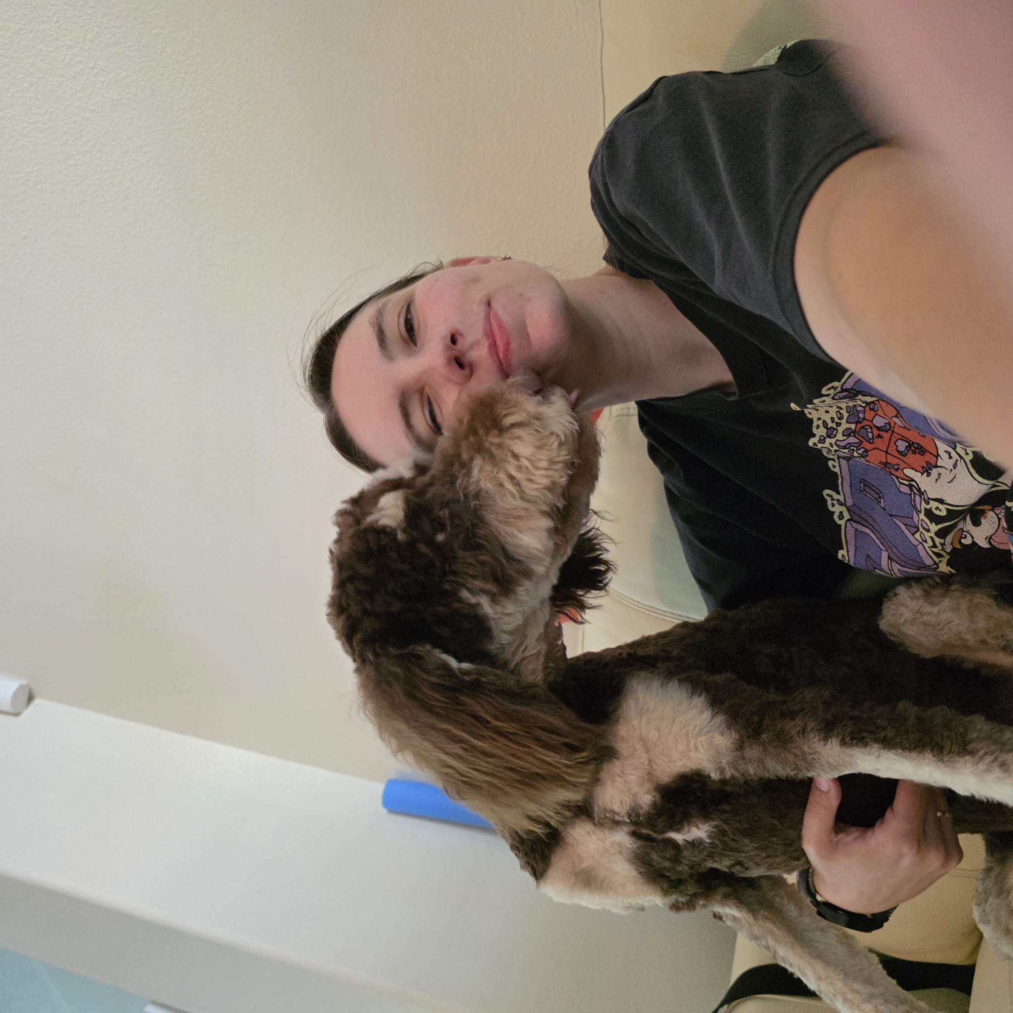 A person is sitting on a couch in what appears to be a living room, holding a dog. The person is looking at the camera with one eye closed and a smile on their face. The dog is a brown and white poodle-mix, and it is leaning in to lick the person's face. The person is wearing a black t-shirt with a graphic print. The couch is beige. In the background, there is a light-colored wall and a blue yoga mat. The scene appears to be indoors, possibly in a residential setting in Portland, Oregon, United States of America.