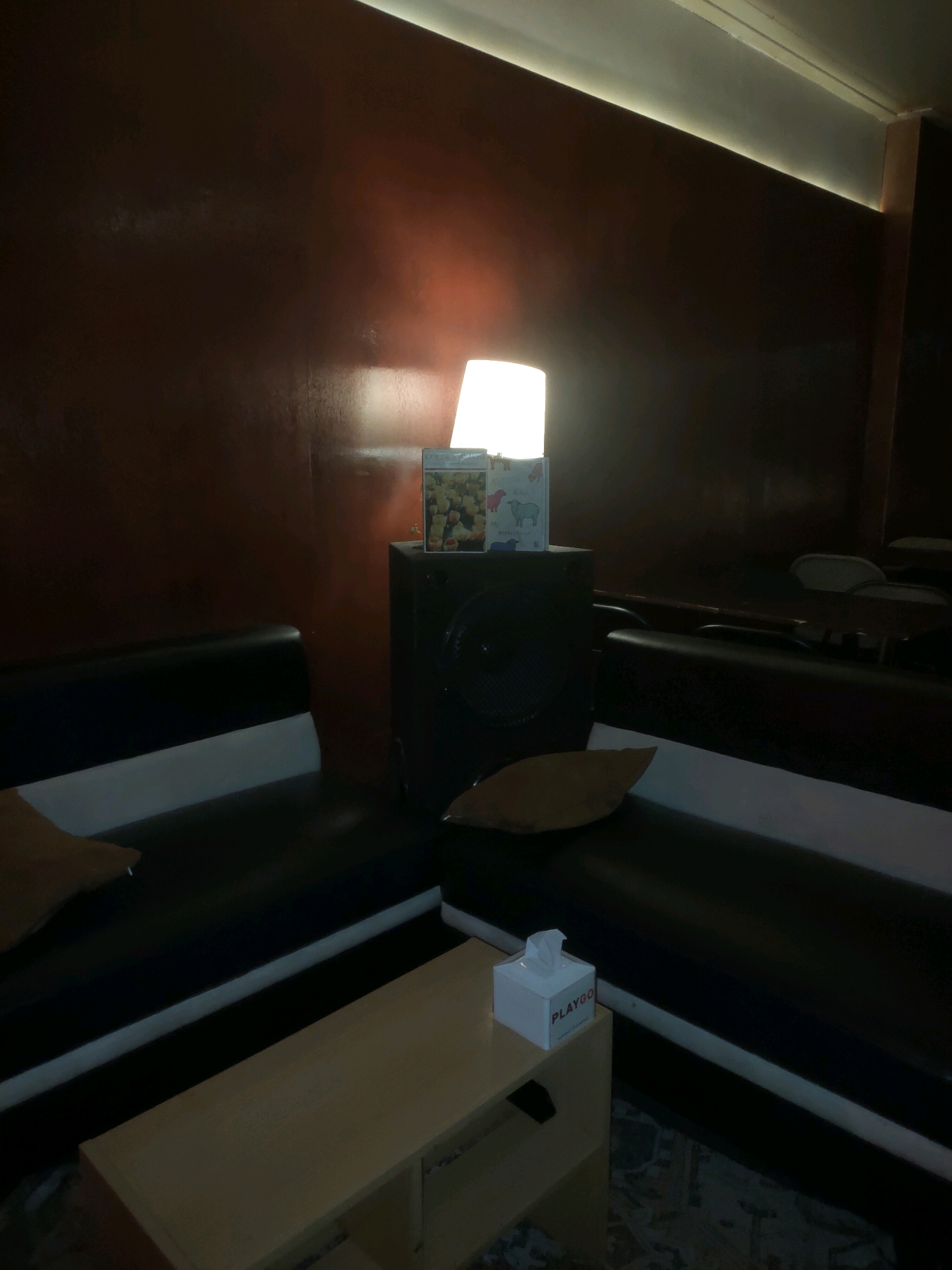 In a dimly lit room, possibly a karaoke booth or lounge in Pagerwojo, Indonesia, a black speaker is positioned on the floor. On top of the speaker, a stack of items is visible: a white lamp with a warm glow, a book with a cover featuring yellow flowers, and a small blue and white box with illustrations of sheep.
The room features a sectional sofa with black upholstery and white accents, arranged in a corner. A light brown throw pillow rests on the left side of the sofa. In the foreground, a light brown coffee table with a simple design is placed in front of the sofa. On the table, a small white tissue box with red lettering that says "PLAYGO" is visible. The walls appear to be a dark, reddish-brown color, and ambient lighting is provided by a light strip along the ceiling. In the background, the dim outlines of other seating arrangements and chairs are discernible.
