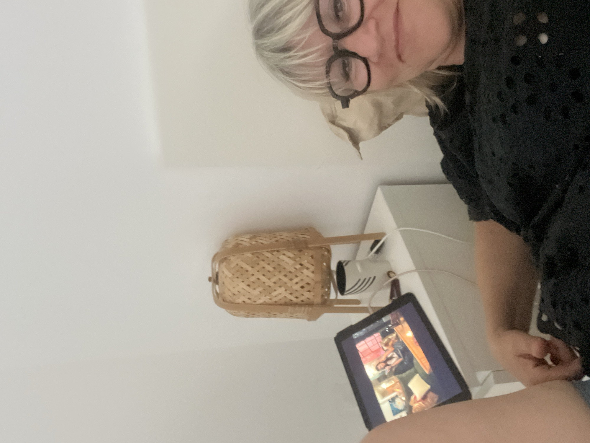 In a bedroom, a woman with blonde hair and glasses is lying on a bed, looking at a tablet computer. She is wearing a black shirt. The tablet is on a nightstand, next to a mug and a lamp with a woven shade. The room is sparsely decorated with a white wall. The image was taken in Chambœuf, France.