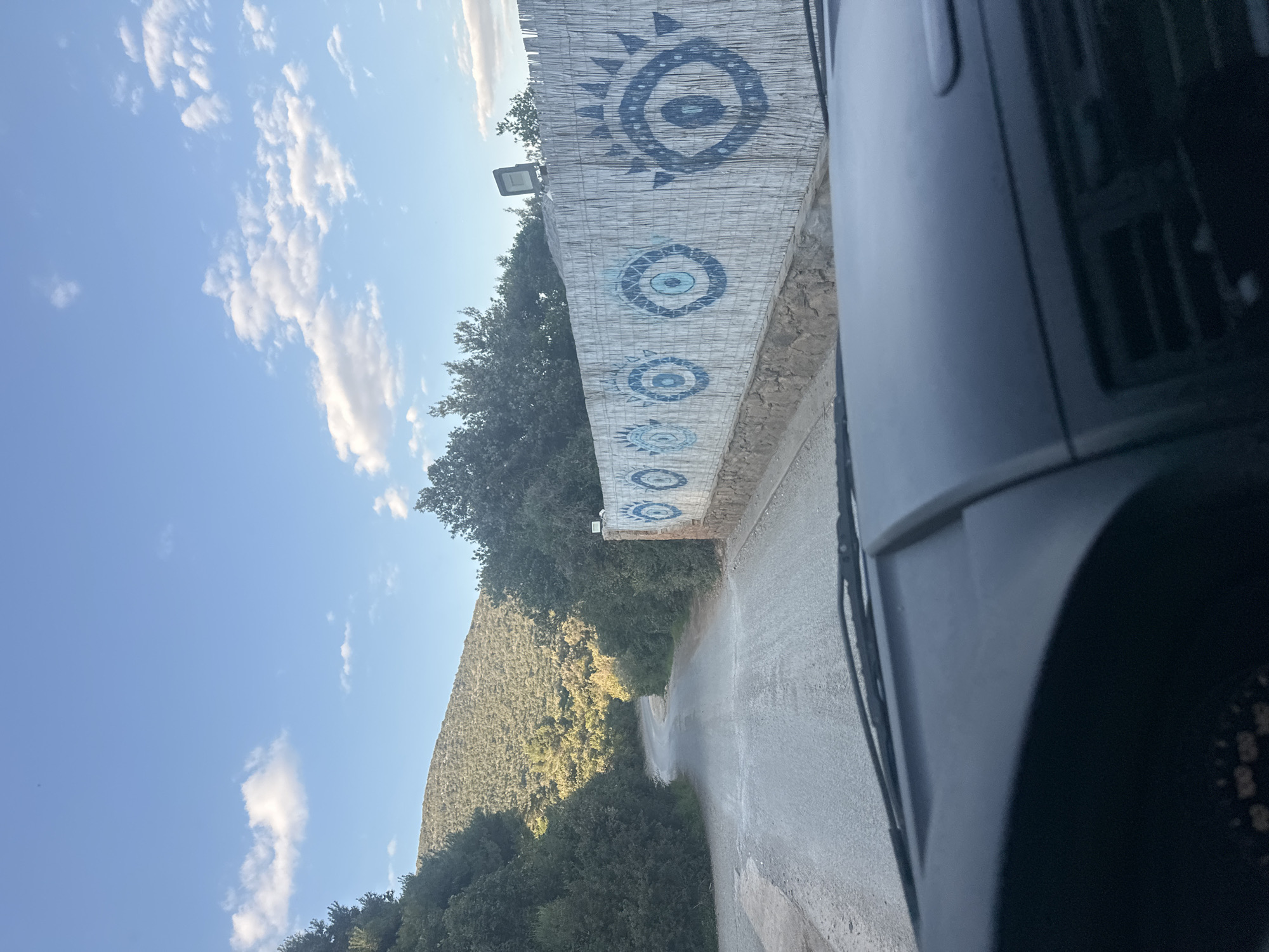 The image shows a view from a vehicle traveling on a road in Gošići, Montenegro. The road is lined with trees and vegetation. A stone wall, decorated with painted blue eye designs, is present on the right side of the road. A hill or mountain covered in trees is visible in the background on the left. The sky is blue with scattered clouds. The foreground of the image contains a portion of the vehicle's interior and exterior.