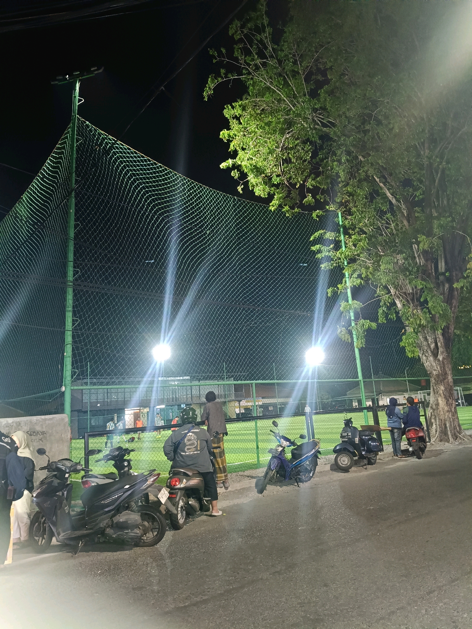 The image depicts an outdoor scene at night, likely in Surabaya, Indonesia, as suggested by the presence of a futsal field and motorcycles common in the region. The focal point is a brightly lit futsal field enclosed by a green net fence. Several floodlights illuminate the field, creating strong beams of light that cut through the darkness.
In the foreground, a row of parked motorcycles is visible on an asphalt road. Two individuals, one wearing a blue jacket and the other a denim jacket, are standing by a motorcycle, seemingly observing the activity on the futsal field. Another person, wearing a grey jacket with a graphic design and a helmet, is seated on a motorcycle. Two other individuals are partially visible on the left side of the frame, one in a white hijab and a dark jacket, and another in a dark hijab and a dark top.
On the futsal field, several individuals are engaged in a game, identifiable by their sportswear. The field appears to be artificial turf. A large tree with lush green foliage is situated to the right of the frame, its branches illuminated by the floodlights. The background is dark, with some building structures faintly visible beyond the futsal field. The overall atmosphere suggests a casual, nighttime gathering around a sports facility.