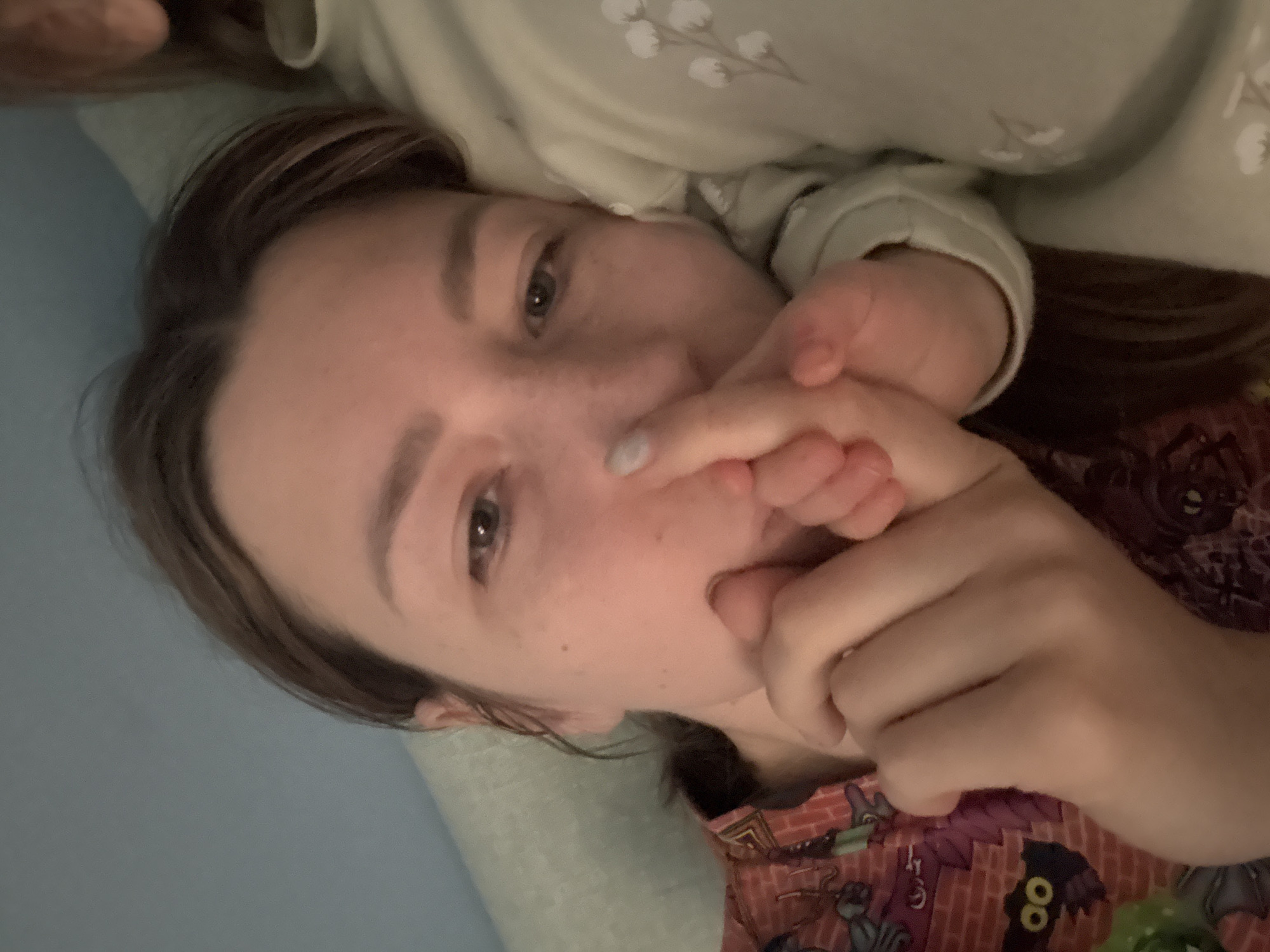 A close-up, angled selfie depicts a young person with light skin, brown hair, and freckles. The person is lying down on a light blue surface, likely a pillow or bedding. Their hand is raised, covering a portion of their face near the mouth and nose. The fingernails appear to be painted a light color. The person is wearing a light green, patterned top, and part of another patterned garment, possibly pajamas, is visible in the lower right corner. The scene appears to be indoors, possibly in a bedroom. There is no indication of the city of Moscow, Russia, in the image.