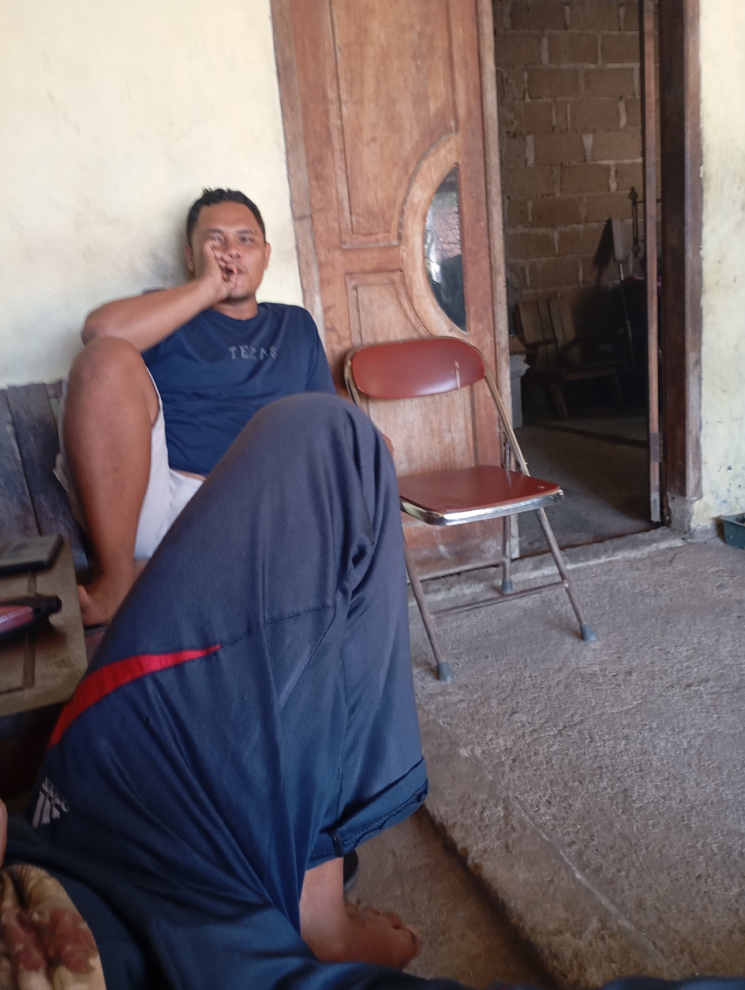 In a low-angle shot taken in the city of Ketepung, Indonesia, a man is seated on a wooden bench against a textured light-colored wall. He wears a dark blue t-shirt with the word "TERAS" in white lettering across the chest and white shorts. His left arm rests on his knee, and his hand is near his mouth, suggesting he may be smoking or in thought. To his right, a reddish-brown folding chair with a metal frame stands on a rough concrete floor. Behind the man and the chair is a wooden door with a circular glass pane, opening into a dimly lit room with exposed brick walls. The foreground of the image is dominated by the legs of another person, wearing dark blue trousers with a red stripe detail. The overall impression is of a casual, indoor or semi-outdoor setting.