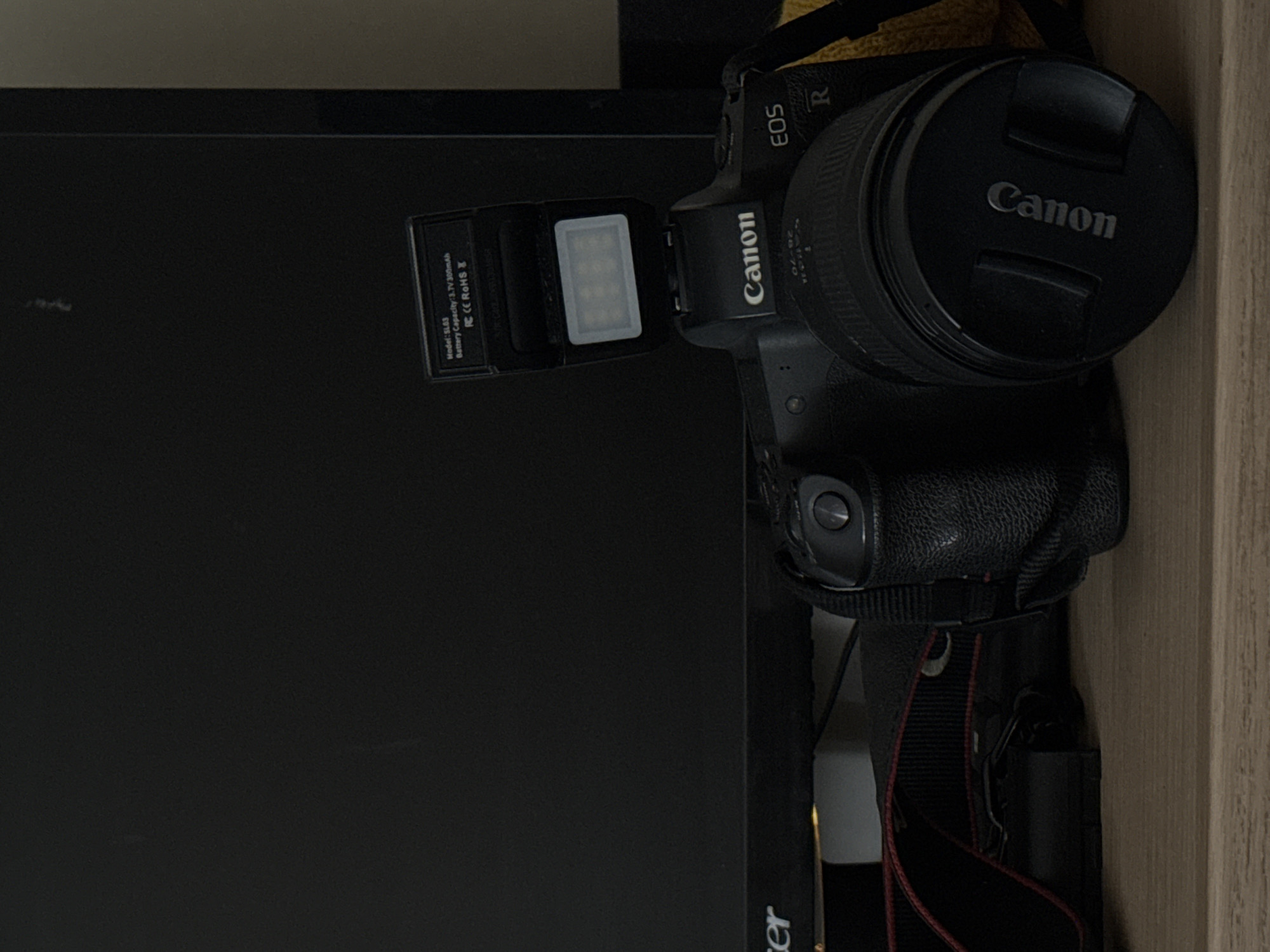 The image shows a Canon EOS R camera with a flash unit attached, resting on a wooden surface in front of a dark monitor. A black camera strap is also visible. The flash unit has text on it indicating "Model: SL63 Battery Capacity: 3.7V 300mAh FC CE ROHS". The background is dark and out of focus. There are no people or animals present. The scene appears to be indoors, possibly a desk or table setup. There is no information in the image to suggest any connection to the city of Kolomna, Russia.
