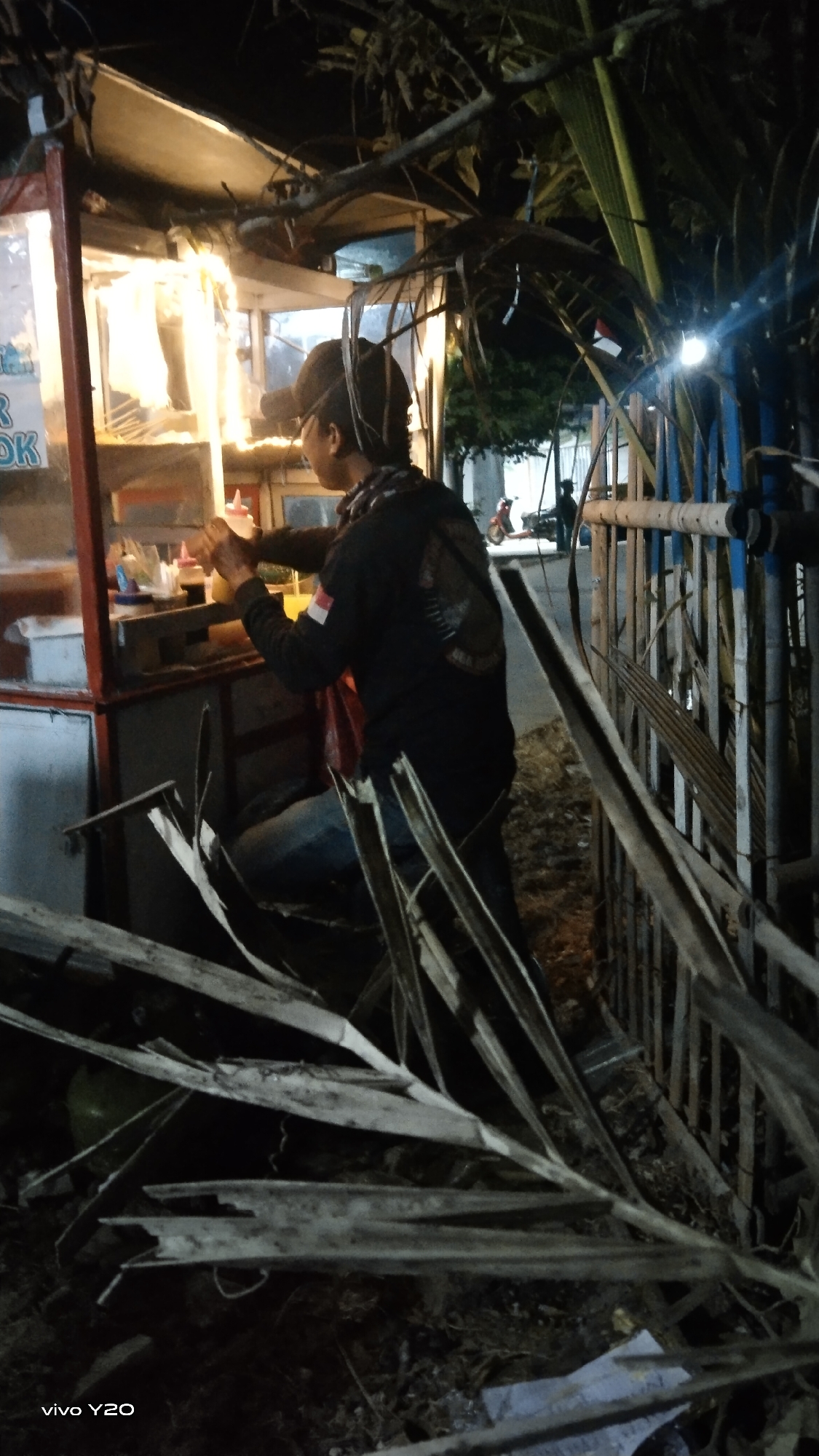 The image captures a nighttime scene in Karawang, Indonesia, likely on a street or in a market area. The foreground is dominated by scattered, broken pieces of palm fronds, suggesting a recent storm or natural debris. In the mid-ground, a food stall is illuminated by bright lights, with its glass display case showing various food items. A person, wearing a dark jacket with a white and red Indonesian flag patch on the sleeve and a baseball cap, is actively preparing food behind the counter. The person is holding a bottle, possibly dispensing a sauce or condiment. To the right of the food stall, a metal fence with blue and white painted sections lines the street. Further in the background, a motorcycle is parked, and another individual is partially visible near it, along with a glimpse of a building or structure. The overall impression is that of a busy, yet somewhat untidy, urban environment at night.