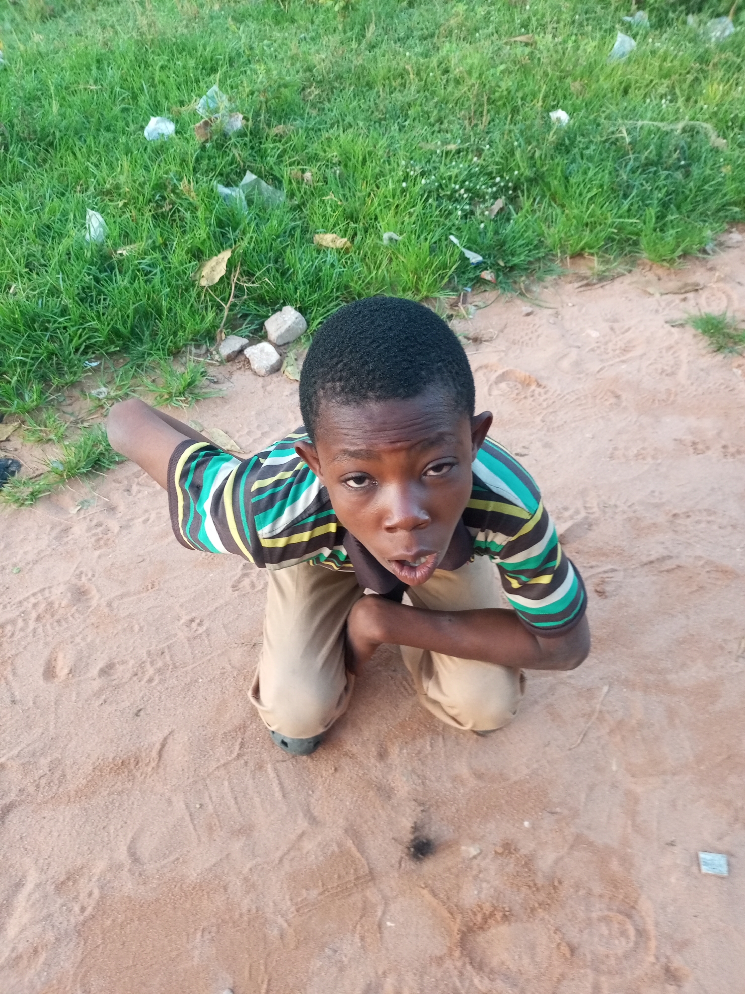 In Tempure, Nigeria, a young boy kneels on sandy ground. He wears a striped t-shirt with horizontal bands of green, yellow, white, and black, and light brown trousers. His gaze is directed upwards, and his mouth is slightly open. The background consists of dry grass with scattered pieces of white plastic trash and a few stones. The ground is a mix of sand and dirt with visible footprints.