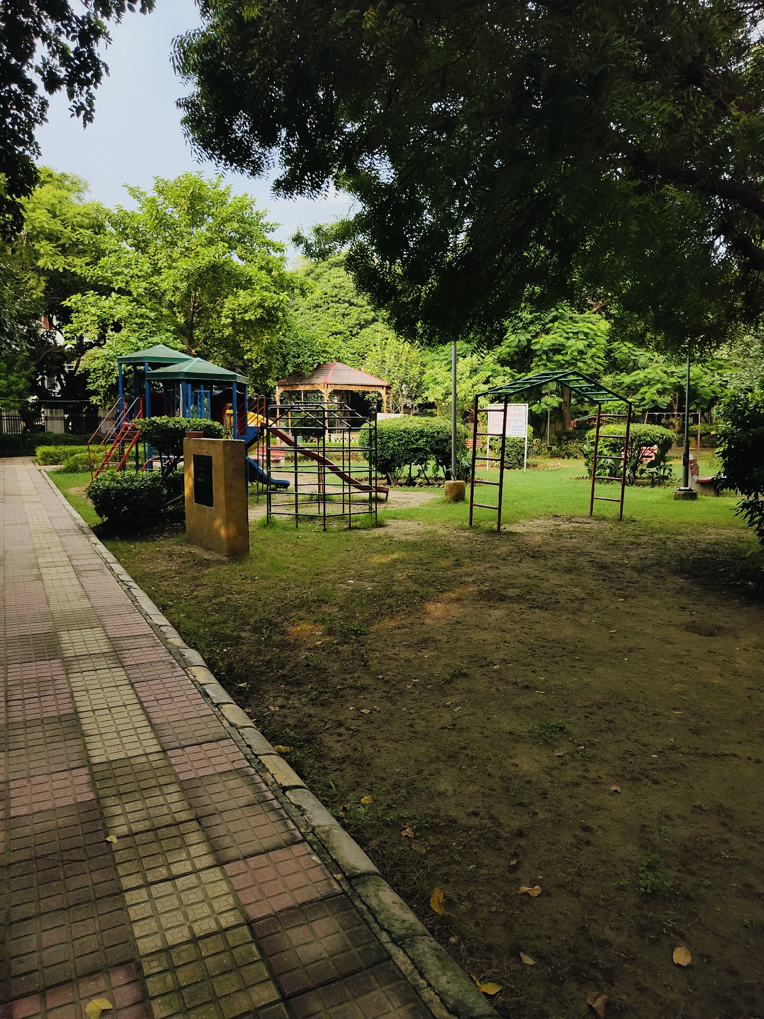The image depicts a public park in Gurgaon, India, featuring a children's play area. A paved walkway with patterned tiles runs along the left side of the frame, bordering a grassy area. In the center, a large, multi-level playground structure with blue and green canopies, slides, and ladders is visible. To the right of this, a metal monkey bar structure with red supports stands on the grass. Further in the background, another covered seating area with a brown roof and an open-sided structure with railings can be seen. The park is surrounded by lush green trees and foliage, suggesting a well-maintained urban green space. The ground in the play area is a mix of dirt and grass, with fallen leaves scattered about. A stone monument with a dark plaque is situated between the walkway and the main playground. The overall scene is peaceful and appears to be empty of people.