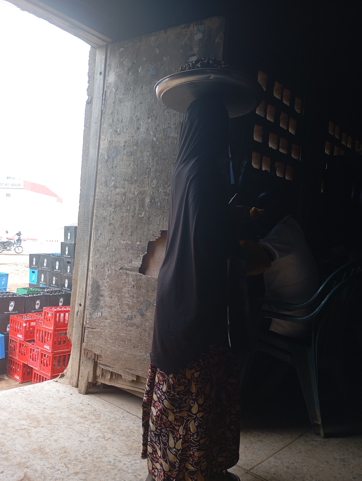 A woman wearing a dark hijab and a patterned skirt balances a large tray of items on her head. She stands near an open doorway, which leads into a dark interior space. Outside the doorway, to her left, stacks of colorful plastic crates are visible, some containing bottles. A motorcycle is seen in the background to the left. Inside the doorway, a blue plastic chair is partially visible in the darkness. The scene appears to be in an outdoor market or storage area.