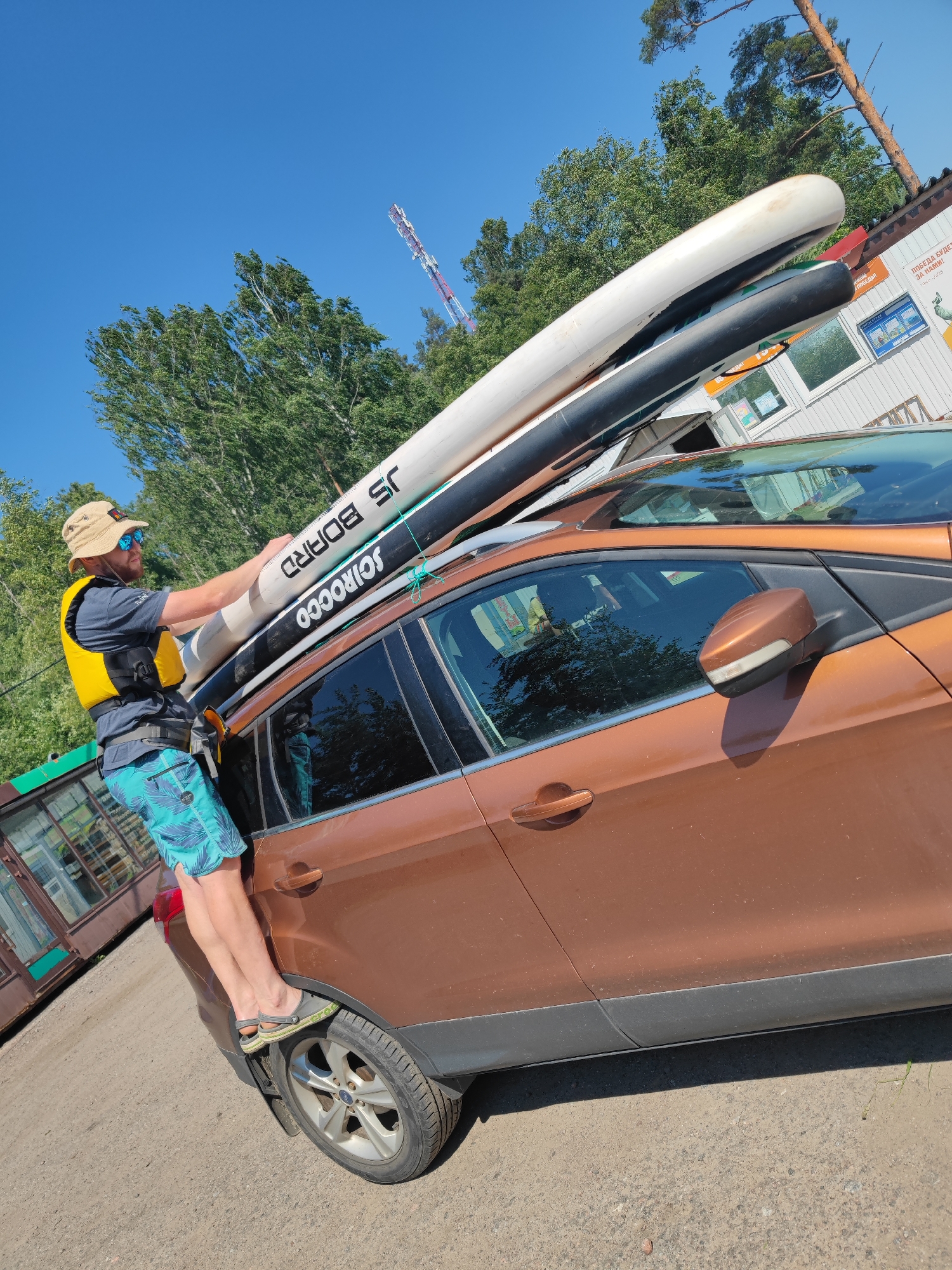 An adult male wearing a straw hat, sunglasses, a yellow life vest, and blue patterned shorts stands on the rear tire of a brown SUV, holding onto two large inflatable paddleboards strapped to the roof rack. The paddleboards are white and black, with "JS BOARD" and "SCIROCCO" printed on them. The SUV is parked on a gravel surface, with trees and a building in the background. A red and white communication tower is visible in the sky above the trees. The environment appears to be a sunny day in a rural or suburban area, possibly near a body of water given the paddleboards. The building in the background has signs in Russian, suggesting the location is in Russia.