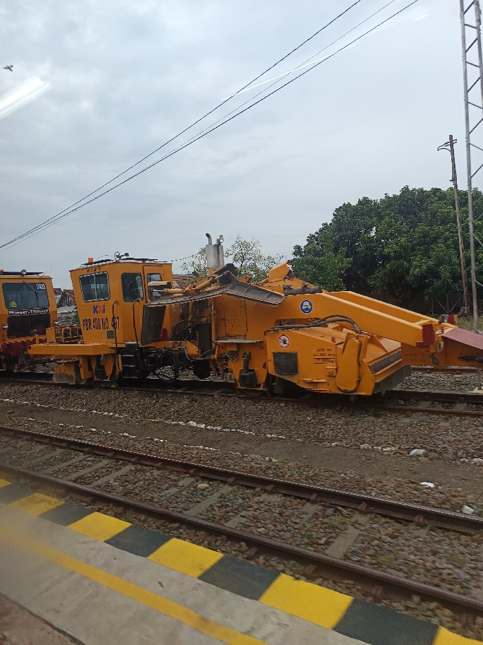 This image depicts two stationary yellow railway track maintenance machines positioned on railway tracks in the city of Probolinggo, Indonesia, under an overcast sky. The primary machine, centrally located, is a complex ballast regulator or track tamper, identified by "KAI PBR 400 NO. 497" painted on its side. It features a control cabin, exhaust stack, and various hydraulic components and brushes for track work. A second, partially visible yellow machine, labeled "PLASSER & THEURER 2420," is coupled behind it to the left. In the foreground, a concrete platform edge with prominent yellow and black striped safety markings is visible. Two sets of railway tracks, laid on a bed of crushed stone ballast, run horizontally across the frame. The background includes dense green foliage from trees on the right, partial structures of distant buildings on the far left, and a metal lattice utility tower on the right. Overhead utility lines traverse the cloudy sky. A small, unidentified light-colored object is faintly visible in the upper left portion of the sky. No personnel are present in the scene.