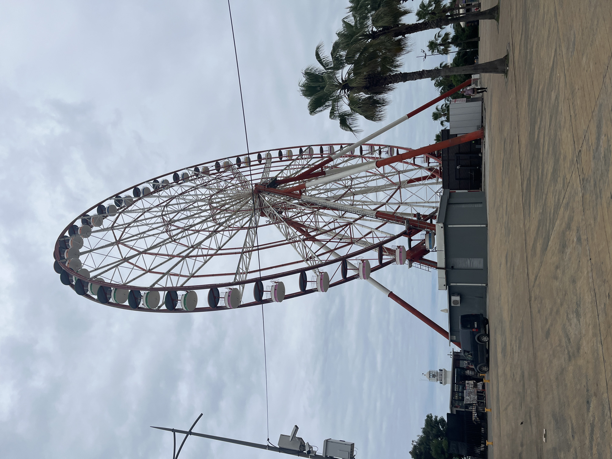 An eye-level, wide shot captures the Batumi Ferris Wheel, a prominent landmark on the Batumi Boulevard in Batumi, Georgia. The towering Ferris wheel dominates the frame, its red and white latticework structure reaching towards a cloudy, overcast sky. Numerous gondolas, alternating in pink and green accents, hang from the wheel's circumference.
At the base of the Ferris wheel, utilitarian grey structures serve as ticket booths or maintenance areas. To the left of these, a black van is parked in a paved plaza. Further in the background, a white lighthouse stands as another visual marker. Several palm trees are positioned to the right of the Ferris wheel, their fronds swaying gently. A lone figure, dressed in a light-colored shirt and dark pants, walks across the plaza to the right. The ground is paved with concrete slabs, marked with shadows from the overcast sky. The overall scene suggests an amusement park or waterfront area, likely during a less busy time of day due to the sparse human presence.