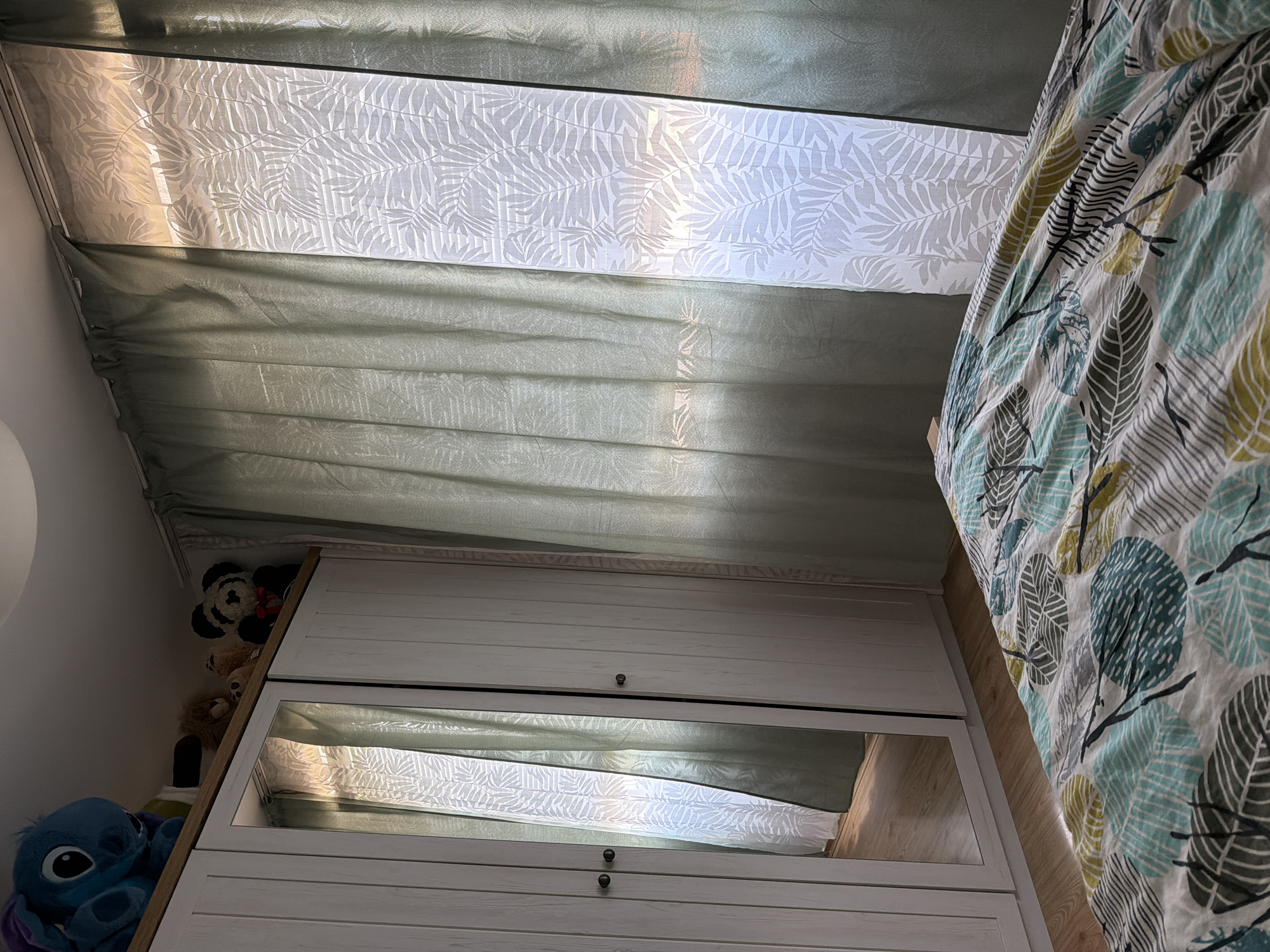 This image captures an indoor scene, specifically a bedroom, located in Plovdiv, Bulgaria. The room is illuminated by natural light entering from a window on the right, which is partially covered by two layers of curtains: an inner translucent white curtain featuring a repeating fern leaf pattern, and outer, more opaque light green drapes. The light source creates a glow through the translucent curtain, highlighting its pattern.
To the left, a light-colored, possibly white or light grey, three-door wardrobe stands on a light wooden floor. The two left doors have circular dark knobs, while the central door is mirrored, reflecting a portion of the room, primarily the patterned white curtain and the floor. On top of the wardrobe, several plush toys are arranged: a large blue character resembling Stitch is on the far left, followed by a brown lion and another smaller light-colored plush toy in the middle, and a black and white panda wearing a red bow tie on the far right.
In the foreground, on the right side of the image, a section of a bed is visible, covered by a duvet or comforter. The bedding features a white base adorned with a distinct pattern of stylized leaves in shades of teal, olive green, and grey, interconnected by dark, thin branches. The ceiling is visible at the top, and a light-colored wooden floor extends from the base of the wardrobe towards the foreground. No individuals are present in the scene.