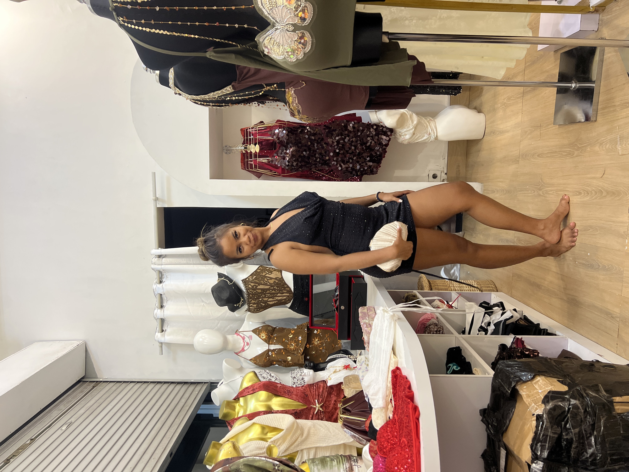 A young woman in a black dress stands on a wooden floor, holding a light-colored clutch, in a room with clothing displays. The room appears to be a boutique or clothing store. The location is in Kuta, Indonesia. Clothing racks and mannequins displaying various garments are visible. The racks are near a window. A white structure with square shelves containing diverse items stands near the woman. A shipping carton wrapped in black plastic sits on the floor.