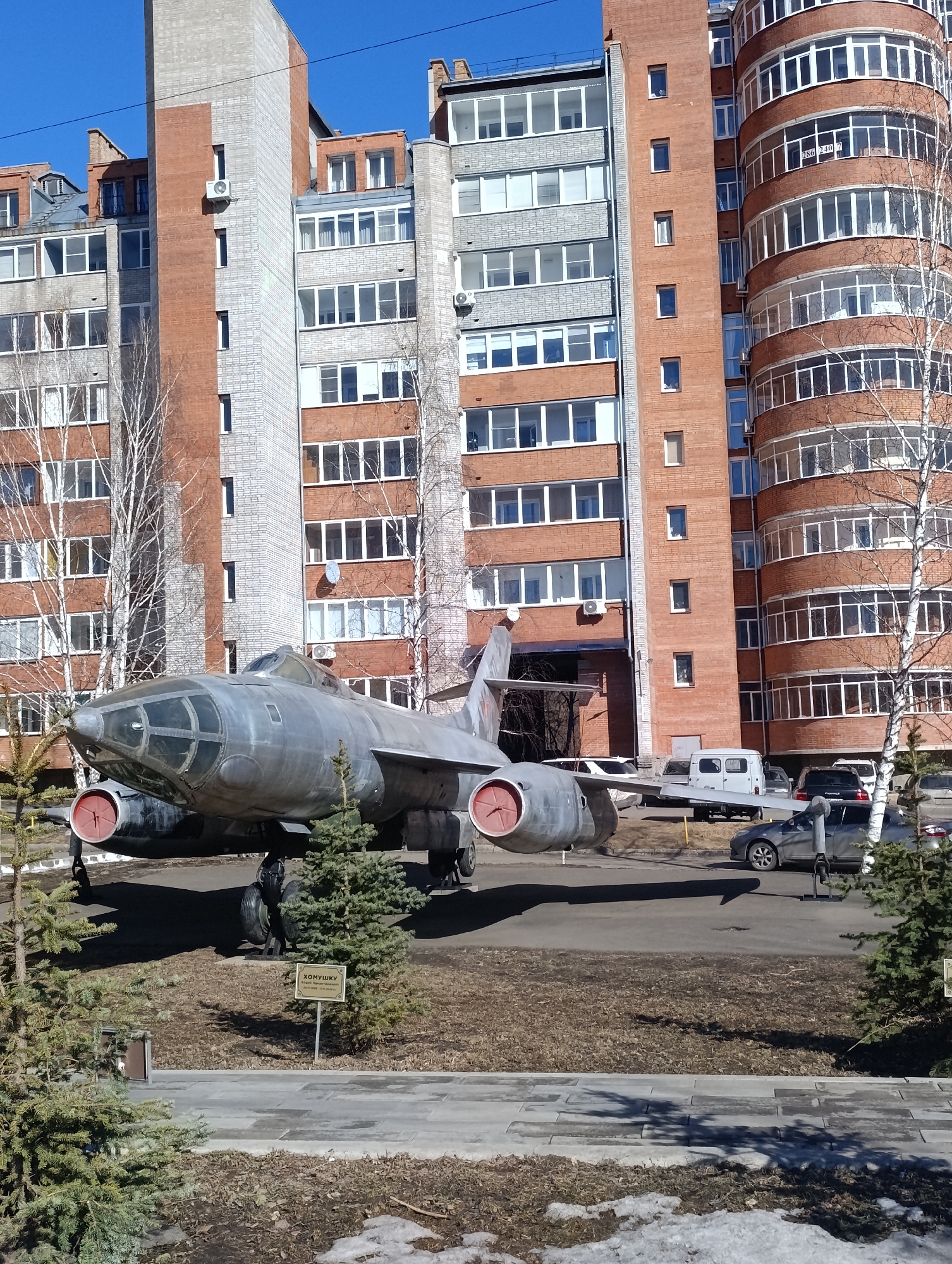 The image depicts a Tupolev Tu-22M (Backfire) strategic bomber on display in Krasnoyarsk, Russia. The aircraft, painted in a faded silver color with red engine inlets, sits on a concrete pad in front of residential buildings. The buildings are primarily constructed of red brick with multiple stories and many windows. Bare birch trees are scattered around the aircraft and the buildings. A small sign written in Russian stands next to the aircraft. Several vehicles are parked in the background. Patches of melting snow are visible on the ground, suggesting early spring. The sky is clear and blue. The overall setting appears to be a public park or monument area within the city.