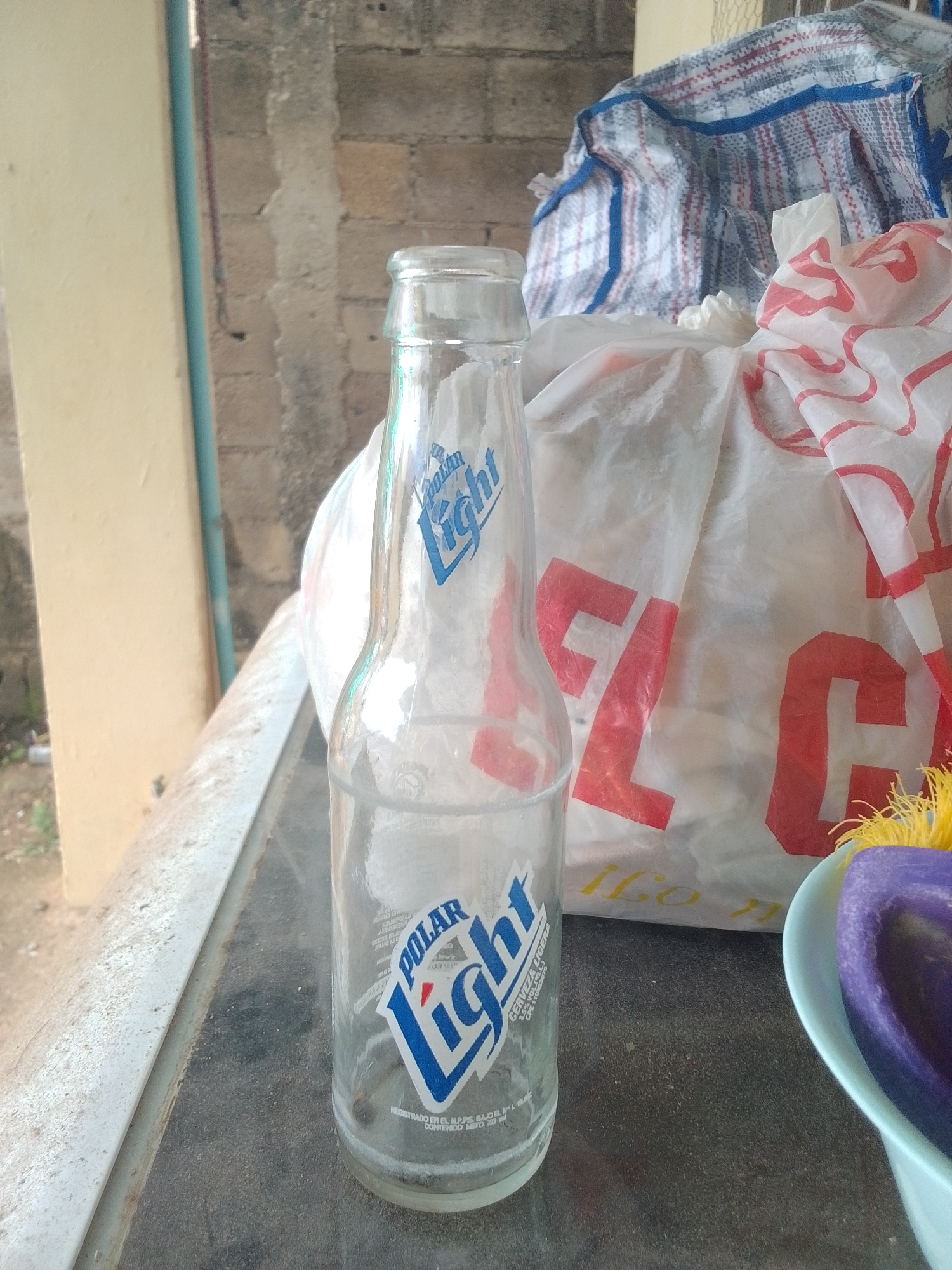 A clear glass bottle with a blue and white label that says "POLAR Light" stands on a dark surface. Behind the bottle, a white plastic bag with red lettering is partially visible, as well as another larger bag with a plaid pattern. The background consists of a textured brick wall and a light-colored vertical structure. The overall scene appears to be outdoors, possibly in a casual setting. There is no visible indication of the time of day or weather conditions. The location is identified as La Tagua, Venezuela.
