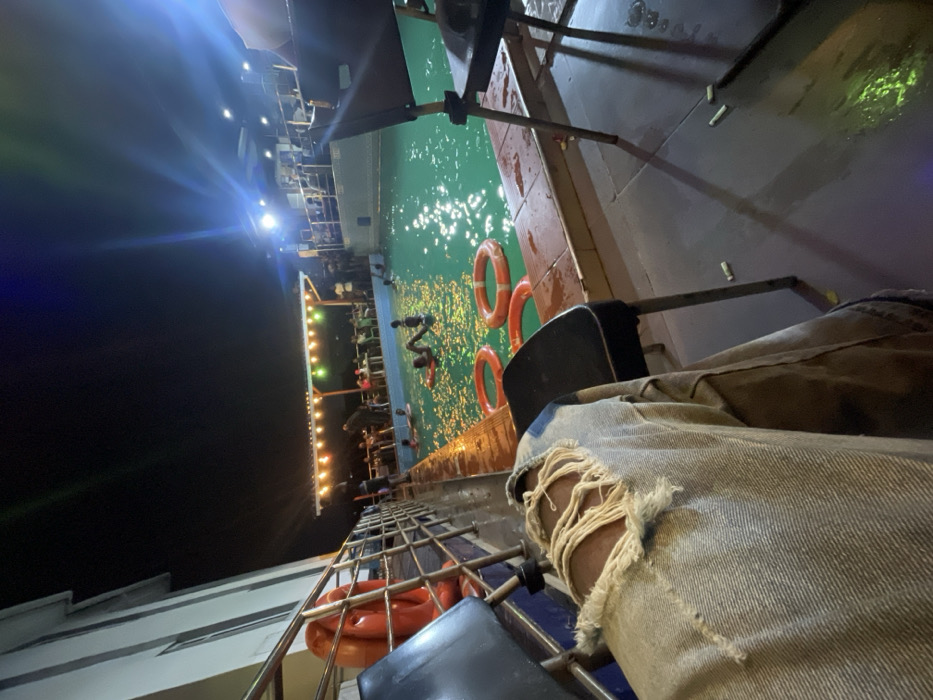 A person sits beside a pool on a boat at night. Ripped denim jeans are visible in the foreground. The pool has several orange life rings, and multiple people swim and stand in the water. The boat is lit with string lights and artificial lights, with part of the deck and upper levels visible. The dark night sky surrounds the boat.
