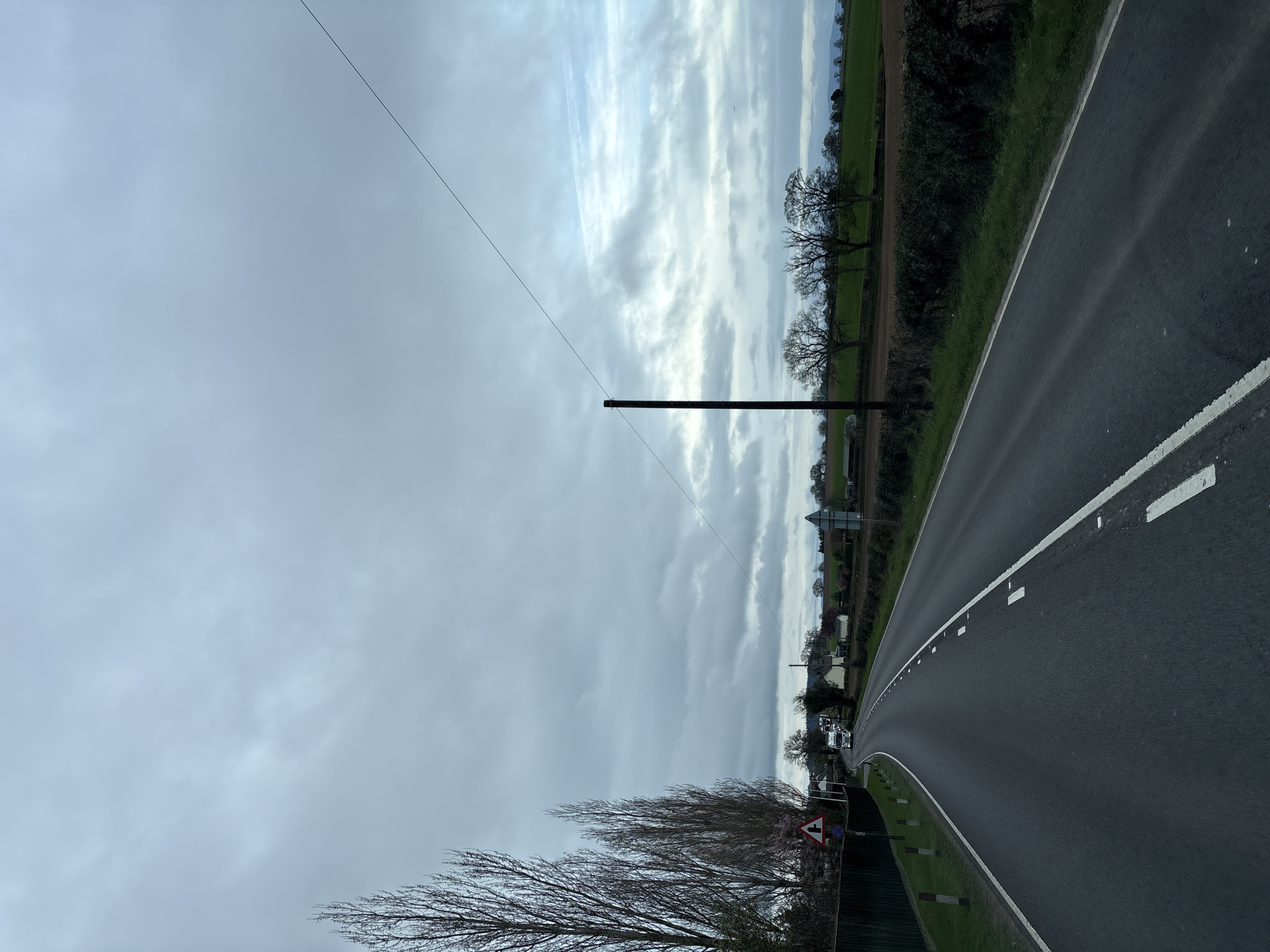 The image displays a two-lane asphalt road under an overcast sky in Ruyton-XI-Towns, United Kingdom. The road curves gently to the left in the foreground and features a solid white line marking the left edge and dashed white lines indicating the center and right edge of the carriageway. On the left, a dark fence runs parallel to the road, succeeded by a row of tall, bare deciduous trees. A red and white triangular road sign, indicating a junction, is visible, along with multiple small white bollards topped with red. Further down the road, faint pink blossoms are present among trees, and several residential buildings are discernible. Two large white vehicles, consistent with lorries or trucks, are observed in the distance, proceeding towards the viewer. On the right side of the road, a green verge borders a dense hedge, beyond which a brown ploughed field and a distinct green field are visible. A prominent dark utility pole stands on the right, from which a single dark wire extends diagonally upwards across the sky. A row of bare deciduous trees lines the distant field edge, and faint hills are visible on the horizon. The sky is characterized by multiple layers of grey overcast clouds with some lighter patches. No individuals are present in the scene.