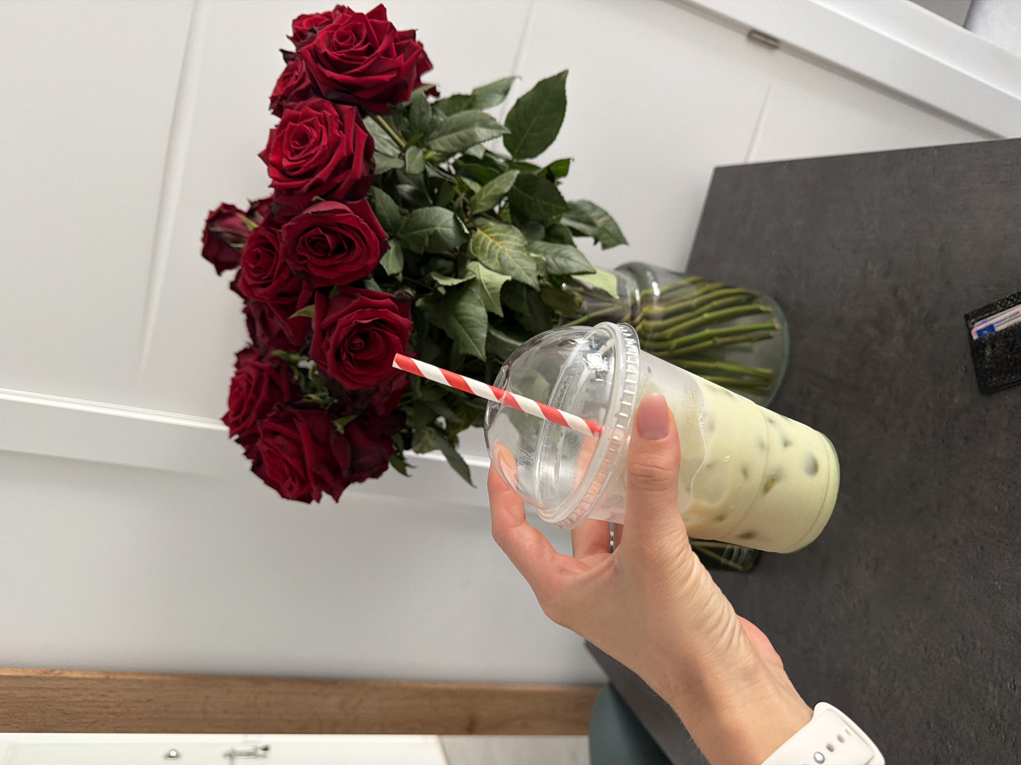 A hand with a white smartwatch is holding a clear plastic cup with a red and white striped straw. The cup contains a pale green, iced beverage with visible ice cubes. In the background, a large bouquet of red roses in a glass vase is arranged on a dark, textured surface. A vertical wooden element is visible to the left, and a white door is to the right. The scene appears to be indoors, possibly a home or a cafe. There is no specific indication of the city of Finestrat, Spain.
