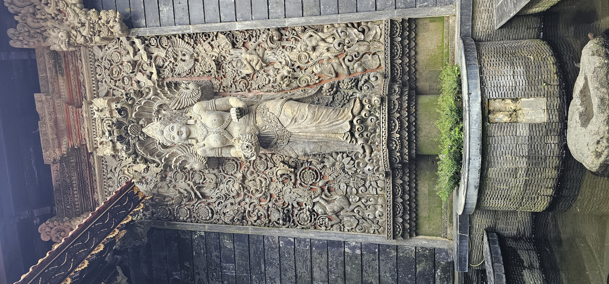 The image captures a detailed stone carving of a reclining figure, possibly a deity, surrounded by intricate floral and avian motifs. The carving is set within a framed, stone-lined structure. Adjacent to the carving is a terraced, semi-circular water feature with vegetation growing along its edges. The overall impression is that this is an example of Balinese architecture, likely part of a temple or shrine. The image was taken in the Ubud District, Indonesia.