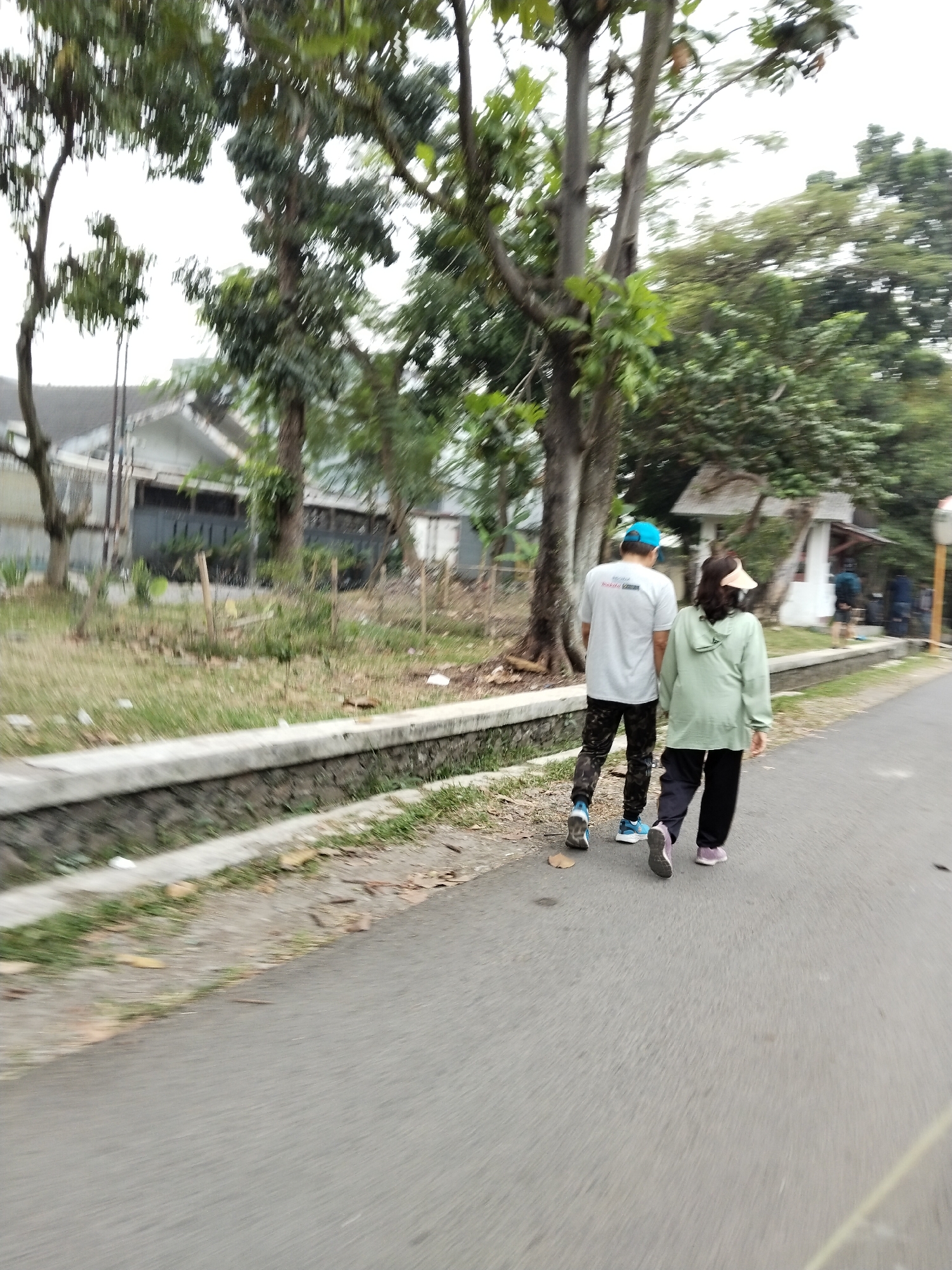 Two individuals, a man and a woman, are walking on an asphalt road in a residential area. The man, wearing a grey t-shirt, black patterned pants, a blue cap, and blue sneakers, walks slightly ahead and to the left. The woman, wearing a light green hooded jacket, black pants, and pink sneakers, walks to his right. They are walking away from the camera. The road is bordered by a concrete drainage ditch on the left, with grass and vegetation growing along its edge. Beyond the ditch, there are houses with white walls and grey roofs, surrounded by trees and foliage. The sky is overcast. The scene appears to be in a suburban or semi-rural part of Bandung City, Indonesia, given the typical housing style and vegetation.