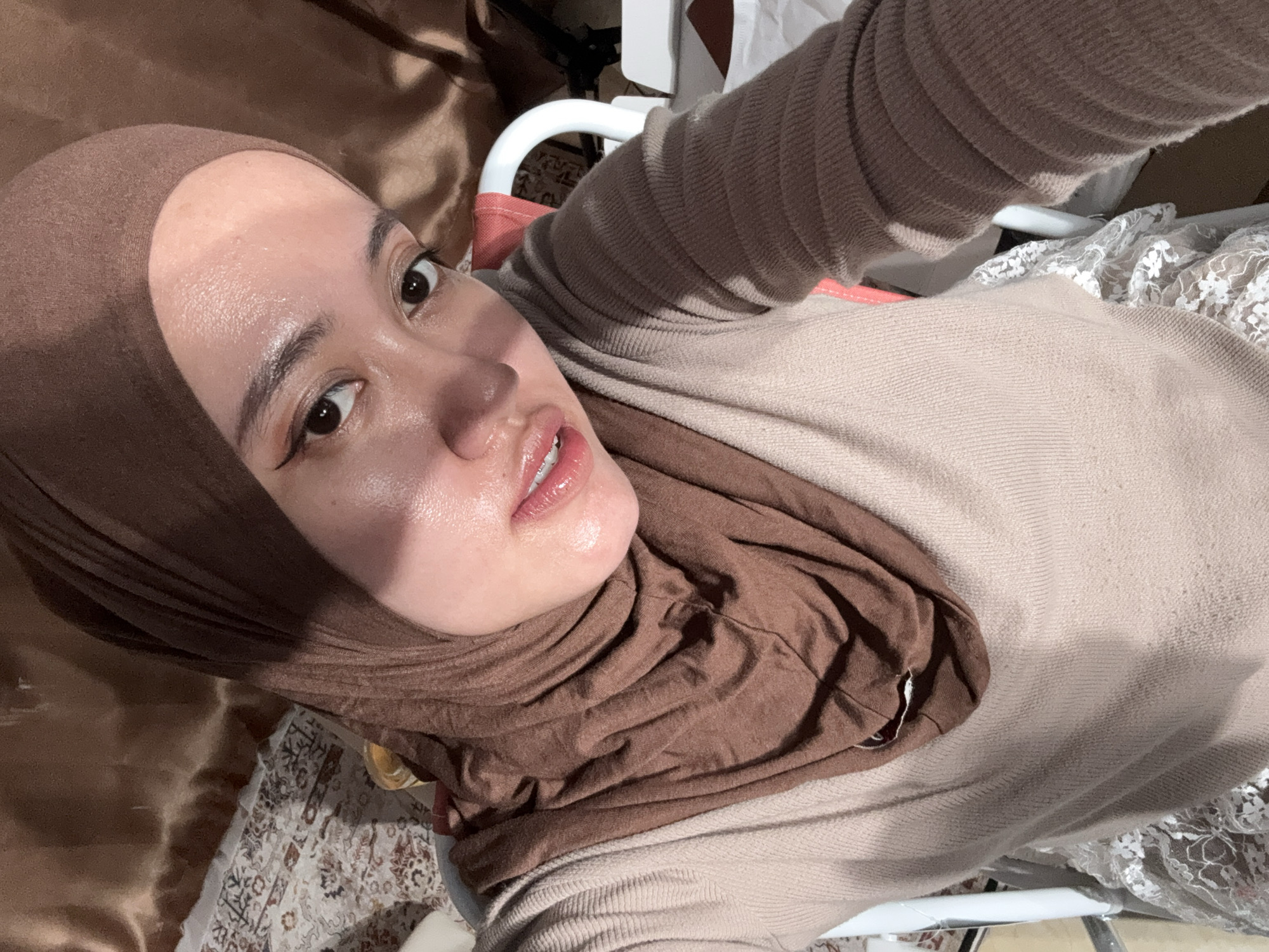 A woman is depicted in a selfie-style photograph. She is wearing a brown hijab and a beige sweater. Her skin appears somewhat shiny, and she has dark eyeliner. The background includes a brown fabric, a white chair, and a patterned rug. The photograph was taken indoors. The location within the city of Gadingrejo, Indonesia is unknown from this image.