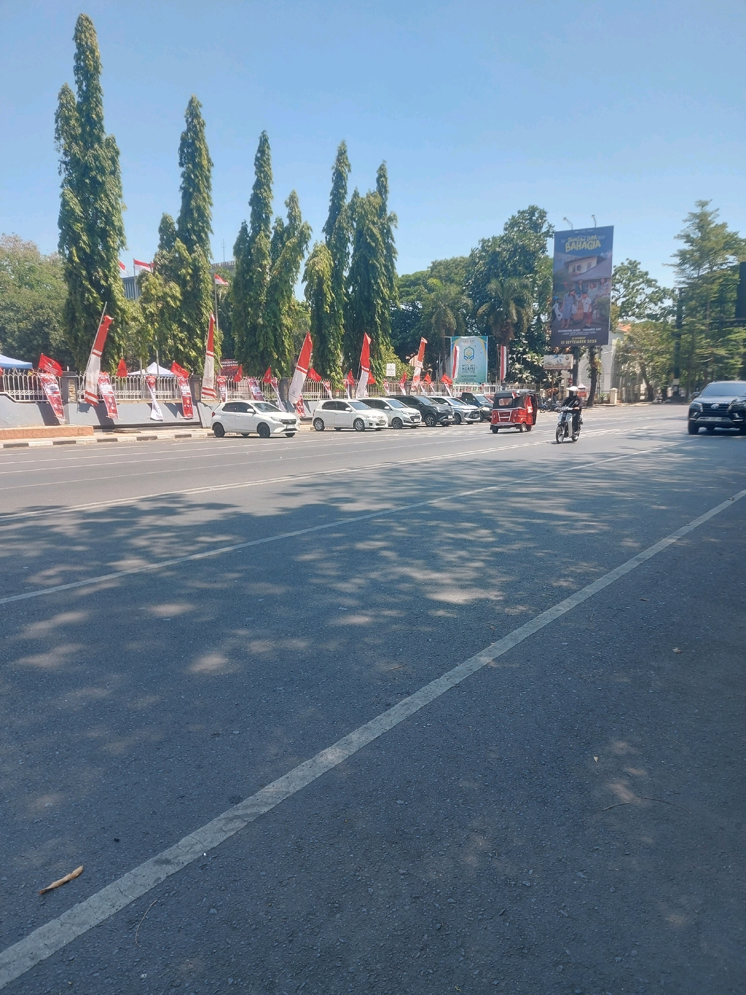 The image depicts a street scene in Makassar, Indonesia, on a clear, sunny day. The foreground is dominated by an asphalt road with a white line dividing the lanes. Shadows of trees are cast across the road, indicating the time of day.
In the background, along the side of the road, is a row of parked cars, predominantly white. Behind the cars, a fence is visible, adorned with numerous red and white flags, suggesting a patriotic or celebratory event. Tall, slender trees with dense green foliage are planted behind the fence.
Further back, a large billboard with colorful images and text in Indonesian is erected. A red three-wheeled vehicle, commonly known as a becak, is visible in the street, along with a person riding a motorcycle. To the far right, a dark-colored SUV is partially visible. The sky is a clear, pale blue. The overall impression is of a well-lit, public urban environment.