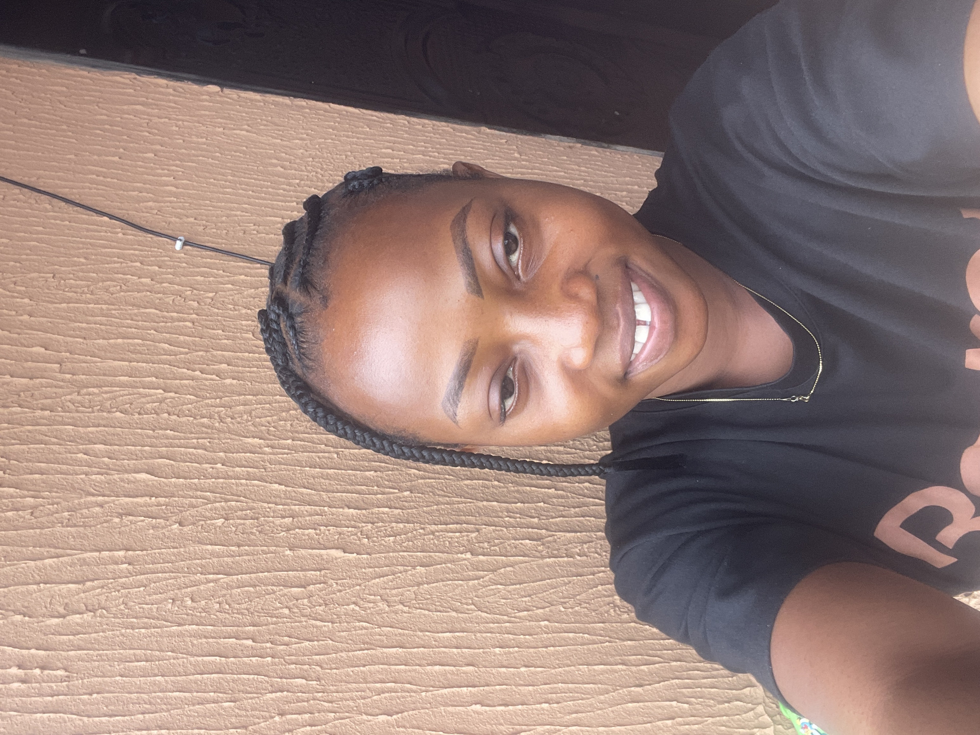 A young Black woman with braided hair and a black t-shirt is shown from the chest up, looking at the camera. She is smiling, with her teeth slightly showing. She is wearing a thin gold chain necklace. She is standing in front of a textured, light brown wall, which appears to be an exterior wall. To her right, a dark doorway is visible. The scene is lit by natural light.