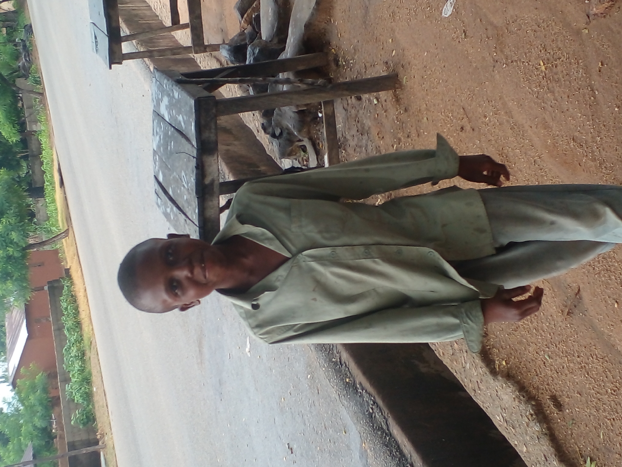 In Langtang, Nigeria, a young person stands on a dirt path next to a road. They are wearing a light green, long-sleeved collared shirt and light gray pants. The shirt is partially unbuttoned, and the sleeves are rolled up. Their right hand is visible, resting near their hip. Behind the person, a weathered wooden table sits near the road, with another similar table further back. The road is paved and appears to be a street in a town, with buildings and trees visible in the background. The ground is a mix of dirt and gravel, with some scattered debris. The overall scene is that of an outdoor environment in a Nigerian town.