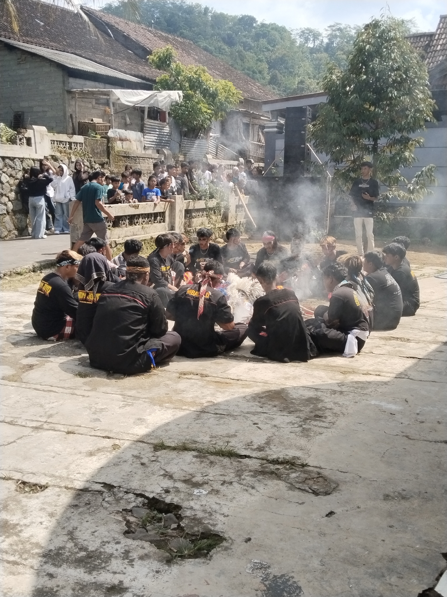 The image depicts a group of people sitting in a circle around a smoke source in an outdoor setting. The location appears to be a village in an unknown city. The central focus is a gathering of approximately 15 individuals seated cross-legged on a concrete surface, seemingly participating in a ritual or ceremony indicated by the smoke and their attire. Many of them are wearing dark shirts with writing on the back and headbands. Behind them, a crowd of onlookers stands near a stone wall, observing the event. In the background are buildings with varying roof structures and green trees, suggesting a rural or semi-rural environment. A large speaker stands nearby. One man stands next to the smoke. The ground shows signs of wear and tear, with cracks and patches.