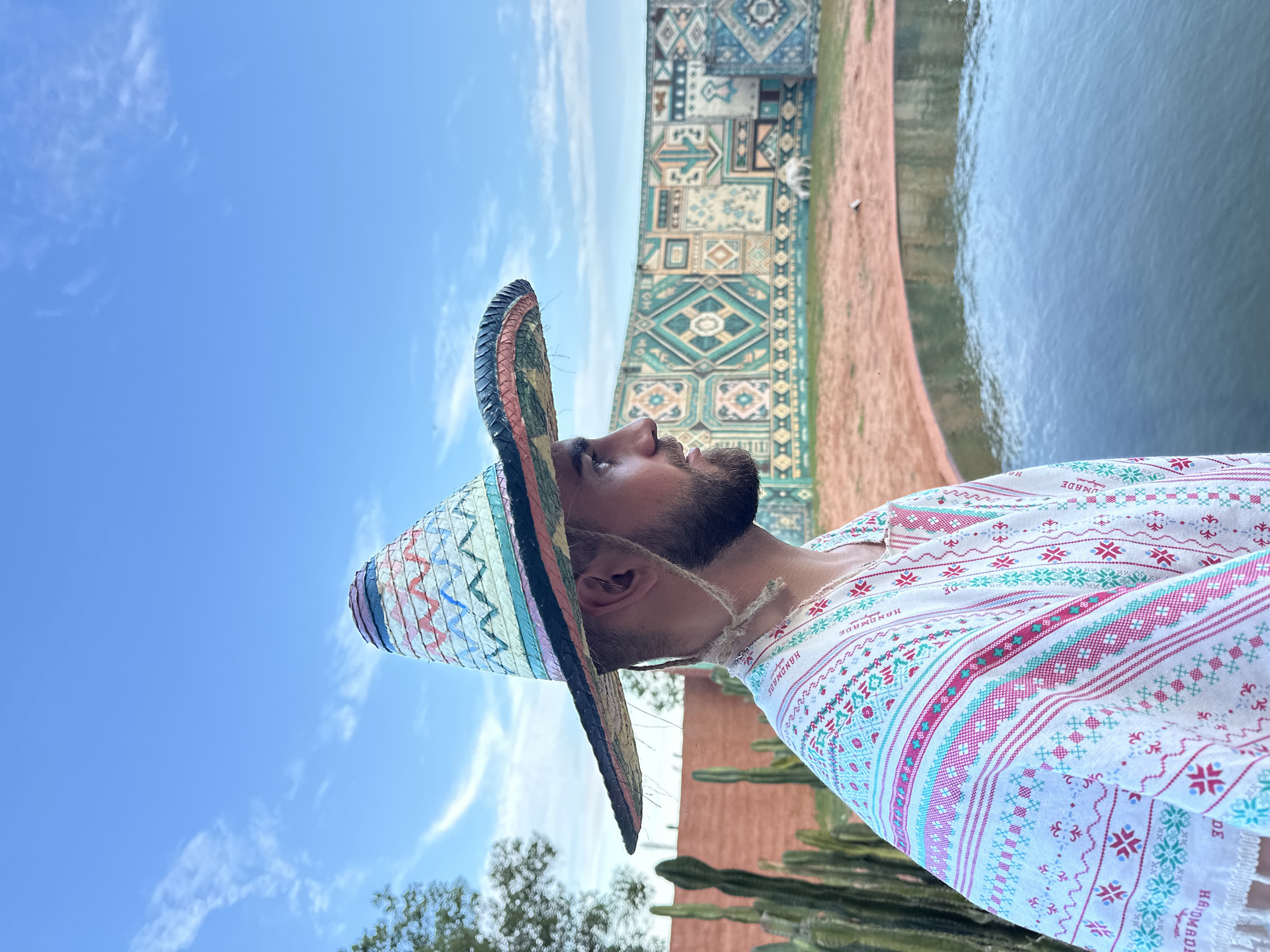 A man wearing a colorful sombrero and a white poncho with pink and turquoise patterns stands in the foreground, facing to the right. Behind him, a decorative wall with geometric patterns is visible, along with a body of water and a few cacti. In the background, a white animal, possibly a goat, grazes. The sky is blue with scattered clouds. The location is likely a resort or tourist attraction in Mui Ne, Vietnam, given the presence of cacti and decorative elements common in such establishments, and the style of the man's attire is reminiscent of traditional Mexican cultural dress, possibly for a themed event or photoshoot.