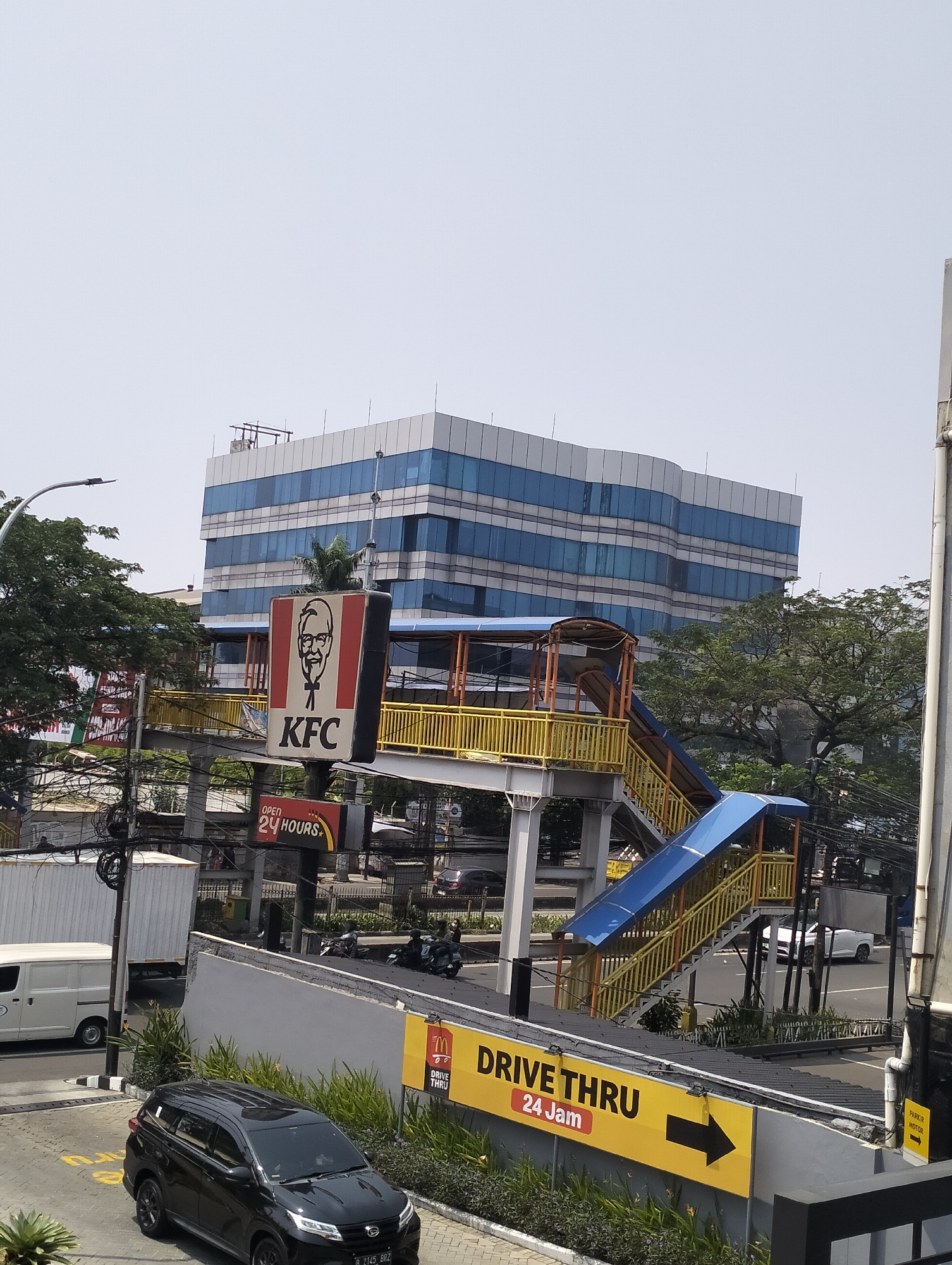 The image captures a street scene in West Jakarta, Indonesia, featuring a KFC restaurant with its iconic signage and a pedestrian overpass. The KFC sign, prominently displayed, shows the founder's portrait and the restaurant's name. Below it, a smaller sign indicates "OPEN 24 HOURS." The pedestrian overpass, painted in yellow and blue, connects two sides of a road, with stairs leading up to it. In the background, a modern, multi-story building with a glass facade dominates the skyline. Trees and utility poles with a tangle of wires are also visible. On the street level, various vehicles are present, including a white van, a black SUV, and several motorcycles. The overall environment suggests a bustling urban setting, common in West Jakarta.