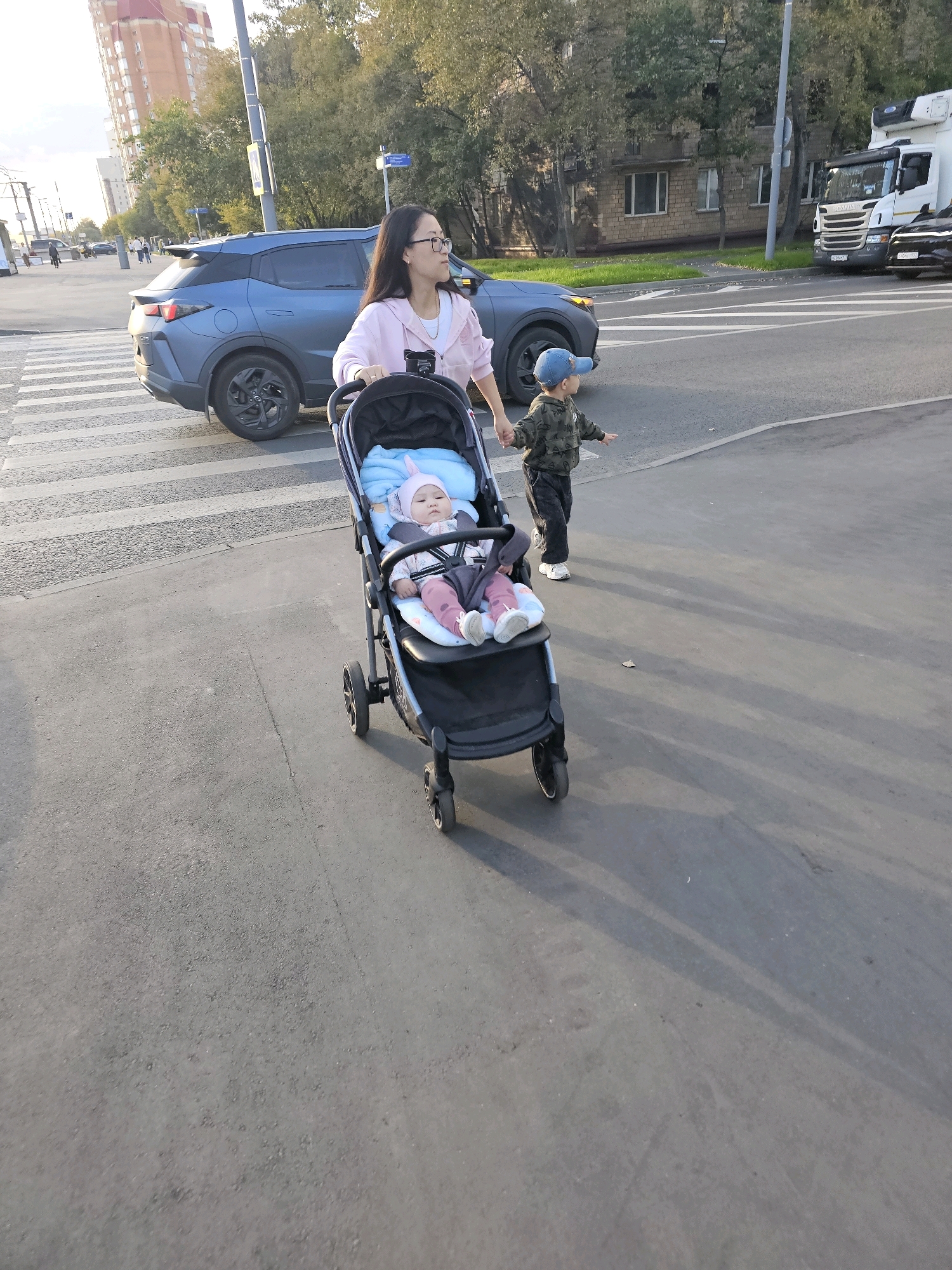 In Moscow, Russia, on a street with a crosswalk, a woman is pushing a stroller with a baby in it. The baby is wearing a pink hat with bunny ears and pink pants. A young boy in a camouflage jacket and blue baseball cap is holding the woman's hand and walking beside her. A blue SUV is stopped at the crosswalk, and a white truck is visible in the background. The scene appears to be taking place during the daytime, with the sun casting long shadows on the asphalt. The surrounding environment includes trees and buildings.