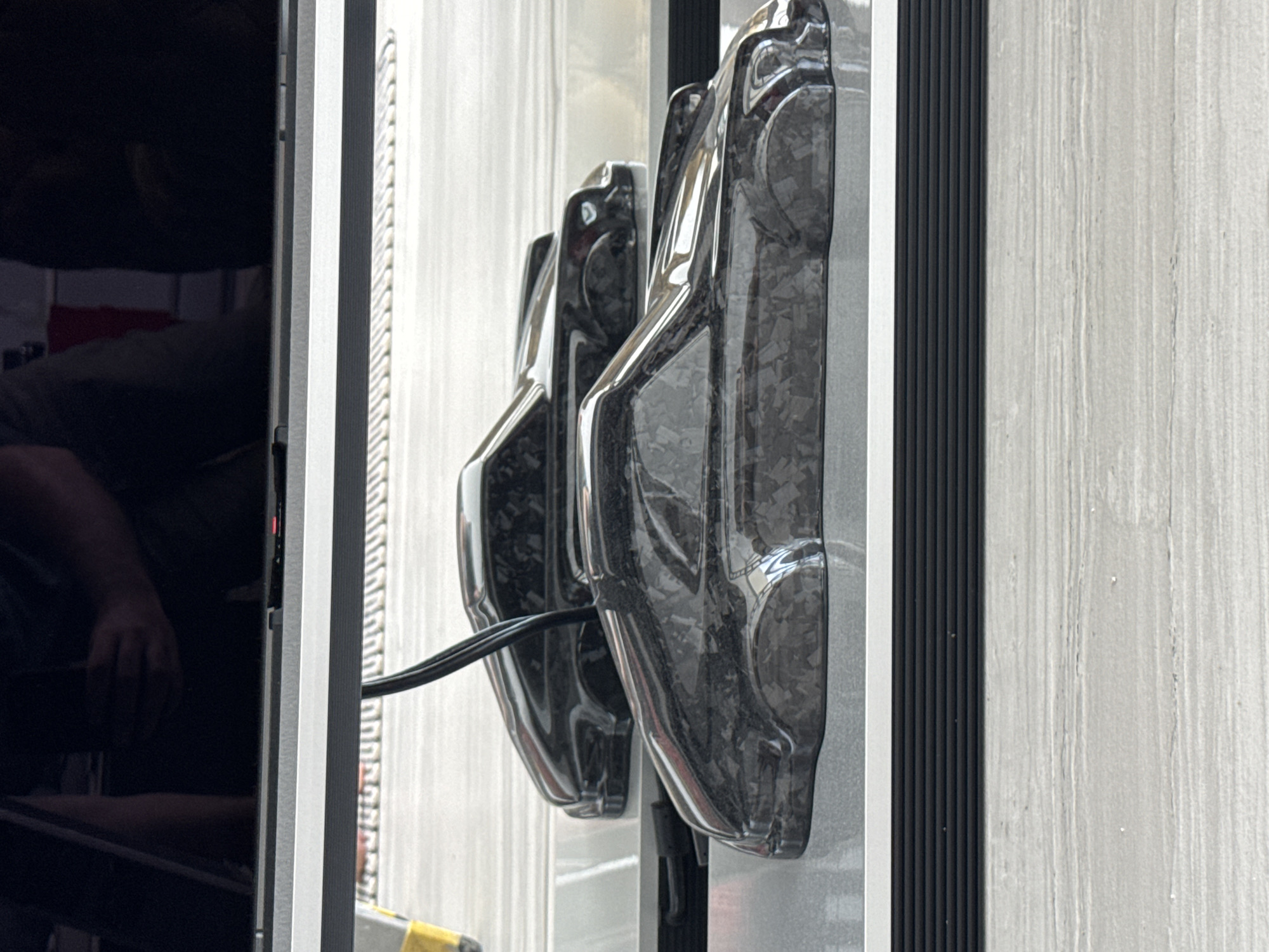 The image shows a close-up view of three dark, reflective objects mounted on a wall. The objects have a curved, angular shape, and their surface is patterned with a mosaic-like texture. A black cable runs between the first and second objects. The wall behind the objects is light gray with vertical lines, and a black strip runs parallel to the mounting. The objects appear to be attached to a frame, which is composed of a white, horizontally-ribbed material and a silver, smooth material. A portion of a dark surface, possibly a television screen, is visible in the background on the left. The location and specific city are undetermined.