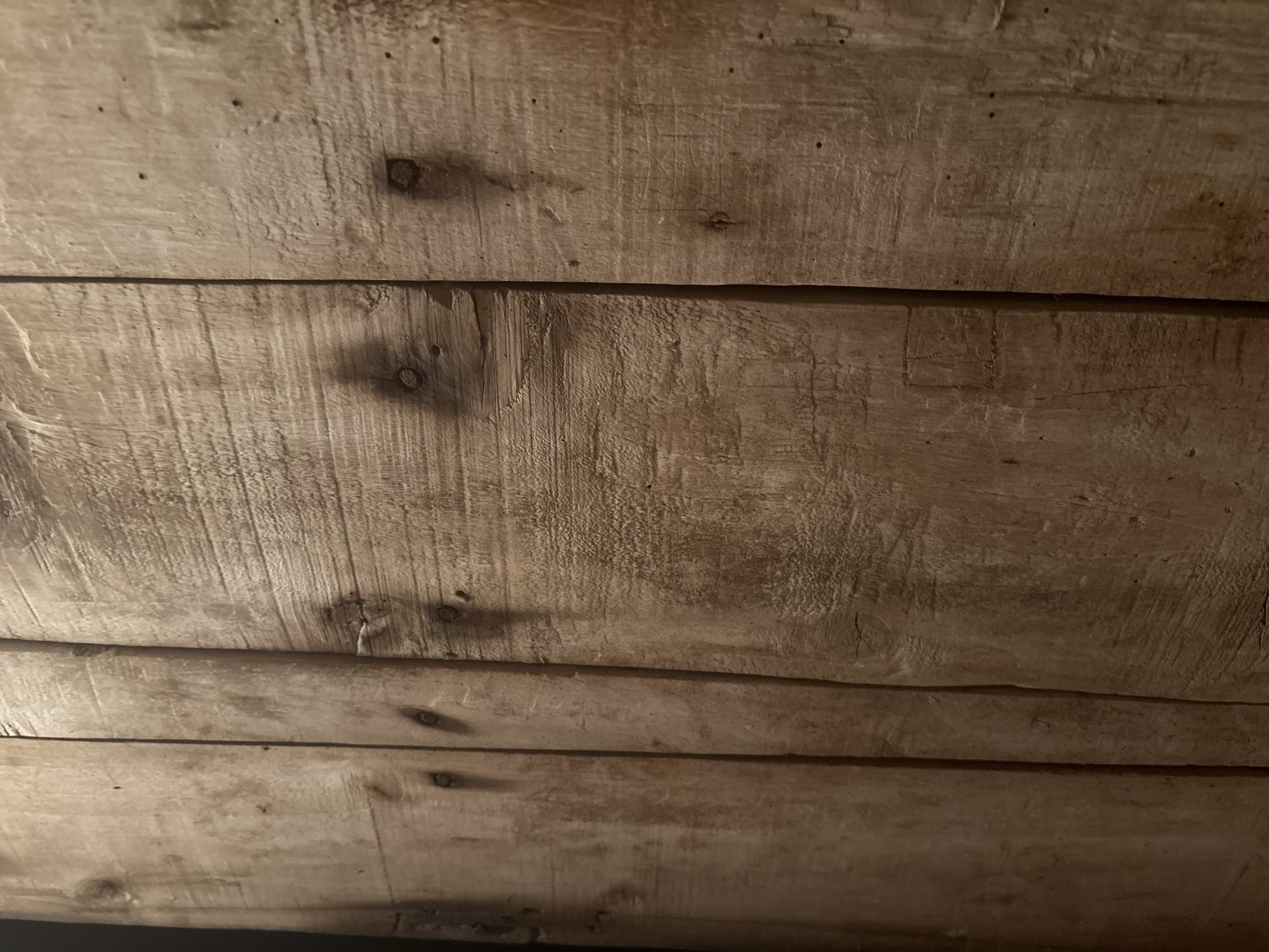 The scene depicts a vertical wooden surface located in the city of Dobrovnitsa, Bulgaria. The surface is composed of multiple light-to-medium brown wooden planks, arranged vertically with distinct seams. The wood exhibits a rough, textured grain, with numerous circular and oval-shaped dark wood knots distributed across the planks. Small, dark, circular indentations, consistent with nail heads or holes, are visible at various points. The environment is dimly lit, with lighting that accentuates the wood's texture and natural variations. No individuals, actions, or interactions are present within the frame.
