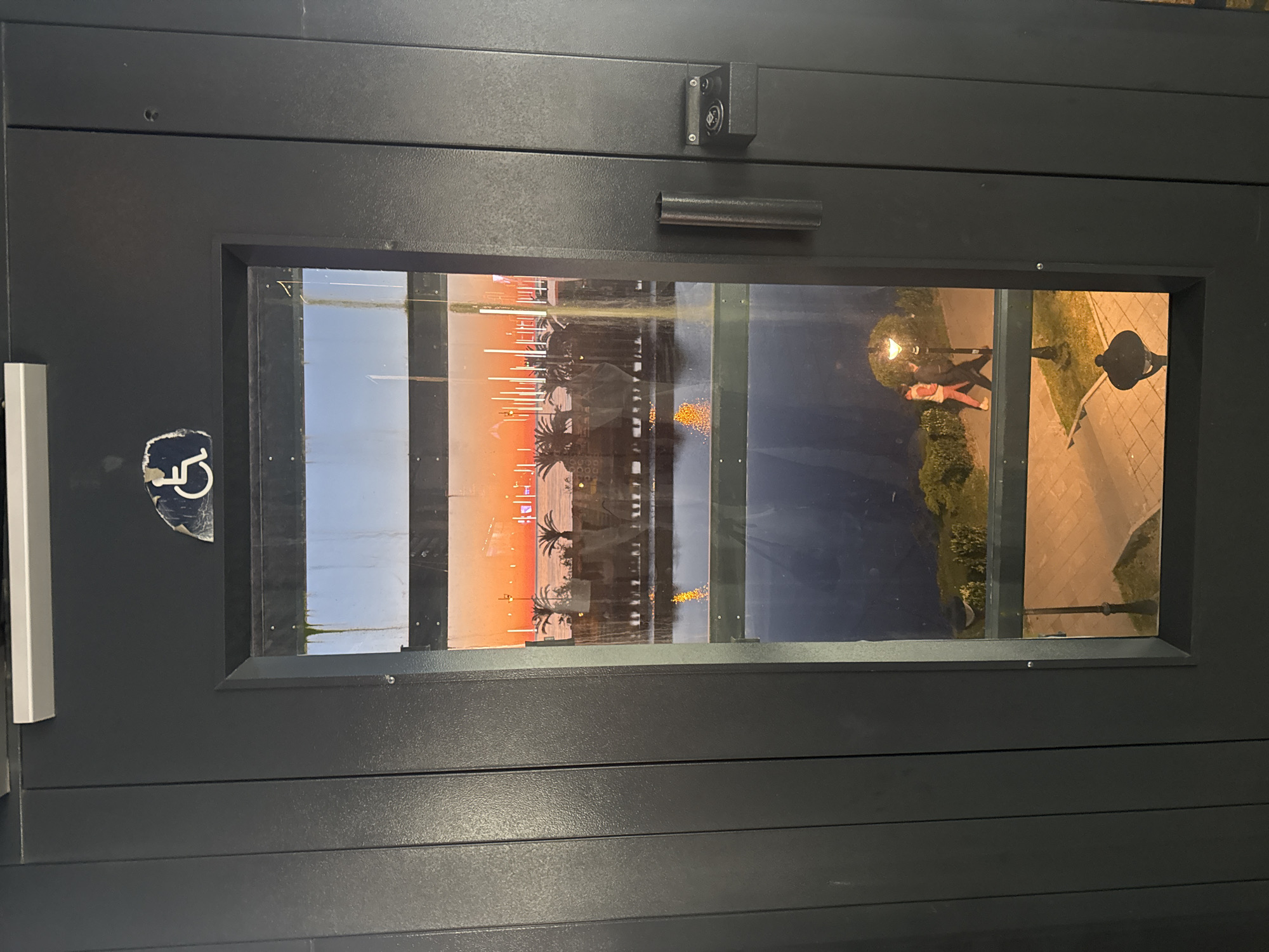 The image shows a close-up view of a dark-colored door with a rectangular window in the center. Inside the window, there is a reflection of a scene in Batumi, Georgia. The reflection includes a waterfront area with palm trees, a sunset sky with orange hues, and a body of water with boats. Additionally, a street scene with people under a lamppost is visible. A sticker with a wheelchair symbol is present on the door to the left of the window. A handle and locking mechanism are visible above the window. The door appears to be part of a building's entrance or access point.