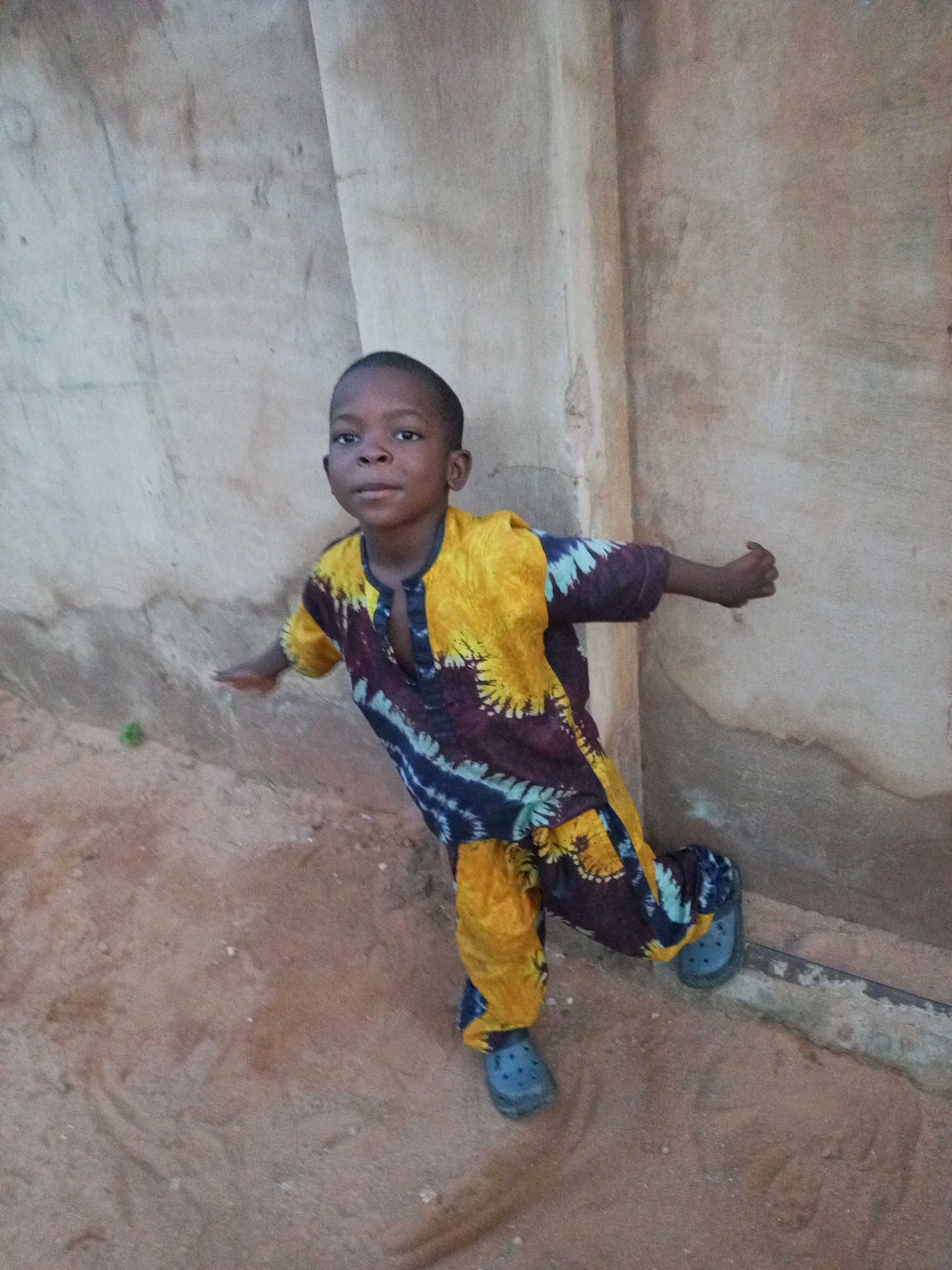 A young child stands in front of a textured wall, on a dirt surface. The child wears a vibrantly colored outfit with a tie-dye pattern in yellow, maroon, and blue. The child's left leg is extended slightly forward and bent at the knee, with a blue sandal on their left foot. The child's right leg is bent and positioned behind, also wearing a blue sandal. The child's arms are outstretched to their sides. The wall behind the child is light brown and has some rough textures and markings. The ground is sandy and uneven, with a few small stones and debris visible. A thin, dark horizontal pipe runs along the base of the wall. The lighting suggests it is daytime, possibly late afternoon. The scene appears to be outdoors, in a simple, perhaps rural or undeveloped, setting. The location is described as Tempure, Nigeria.