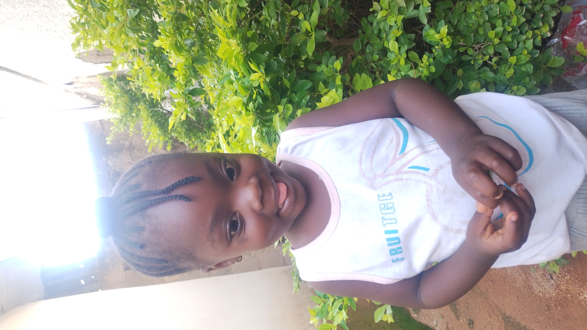 A young African child stands outdoors in a grassy area, appearing to be in a yard. The child is light-skinned and wearing a light-colored sleeveless shirt. The child has dark hair in braids. Behind the child, lush green bushes or trees fill the background. The scene appears to be in daylight, and bright light is visible to the left side of the child, potentially indicating the position of the sun. It is not possible to determine the specific location in the city of Jos, Nigeria from the given information.