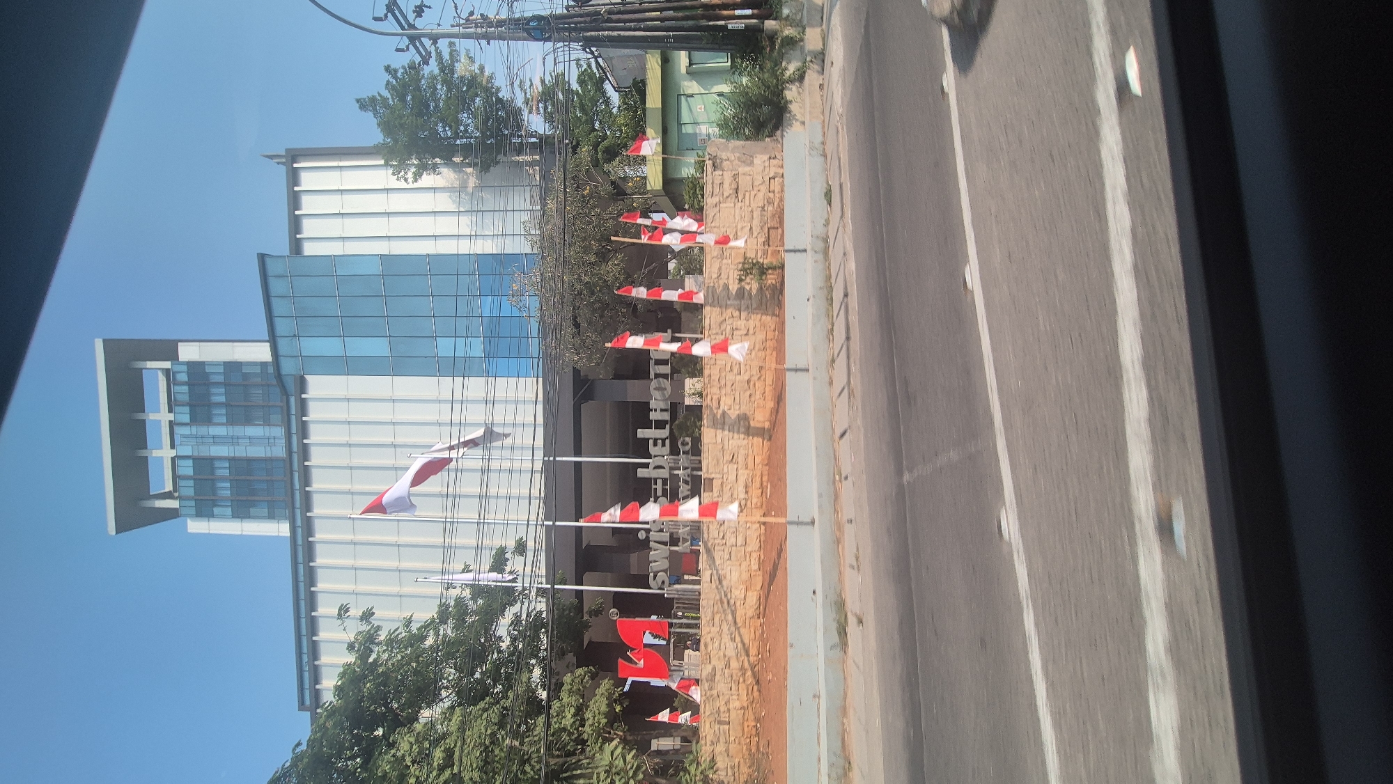 The image captures a modern building with the signage "SWISS-BELHOTEL KARAWANG" prominently displayed at its entrance. The hotel features a distinctive facade with large blue glass windows and white vertical elements. In front of the hotel, a low stone wall separates the property from a paved area, which is lined with numerous small Indonesian flags, alternating red and white stripes. The flags are attached to thin poles and are positioned along the edge of the property facing a road. Trees with green foliage are visible around the building and along the property line. The sky is clear and blue, indicating a sunny day. The image appears to be taken from a vehicle moving along the road, as suggested by the blurred foreground and the angle of the shot.