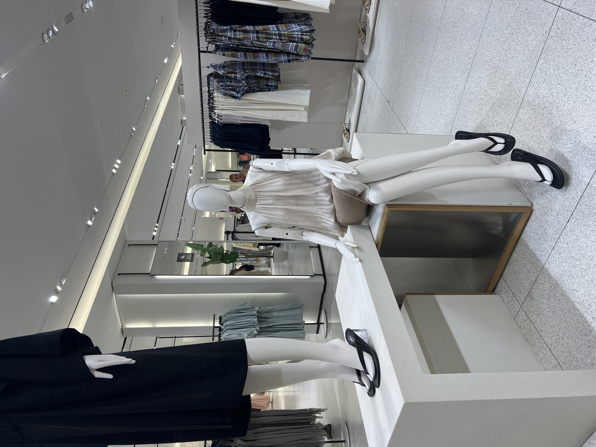 A retail store in Beşiktaş, Turkey, features mannequins displaying clothing and footwear. A mannequin on the left is dressed in a black dress and black flip-flops with clear heels, standing on a white platform. To the right, a seated mannequin wears a light-colored, sleeveless top with a gathered neckline and a neutral-colored skirt, also accessorized with black flip-flops. In the background, clothing racks display various garments, including patterned shirts and solid-colored items. The store's interior has a modern design with white walls, a light-colored tiled floor, and integrated lighting.