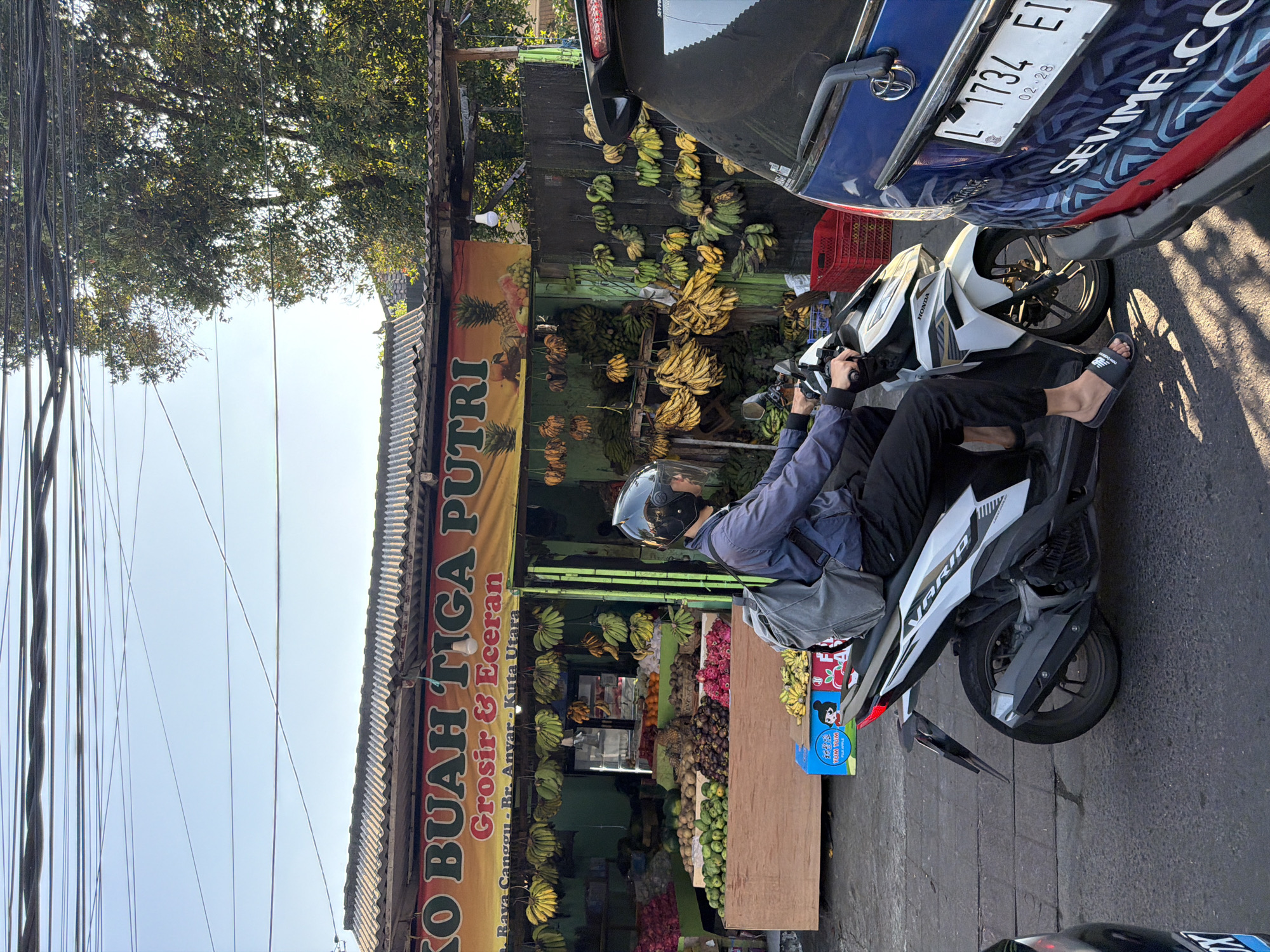 In North Kuta, Indonesia, a person wearing a blue jacket and black pants rides a white and black Honda Vario motorcycle. The rider has a black helmet with a clear visor and is carrying a gray backpack. They are positioned on the right side of the frame, moving forward. To the right of the motorcycle is the rear of a blue vehicle, the license plate reads "L 1734 EI" with the expiry date "02-28". In the background, a fruit stand is visible, displaying an abundance of bananas, pineapples, and other fruits. The fruit stand has a yellow banner with the text "KO BUAH TIGA PUTRI Grosir & Eceran" and an address "Raya Canggu - Br. Anyar - Kuta Utara". The scene is illuminated by natural sunlight, creating shadows on the road. The environment appears to be a street in a city.