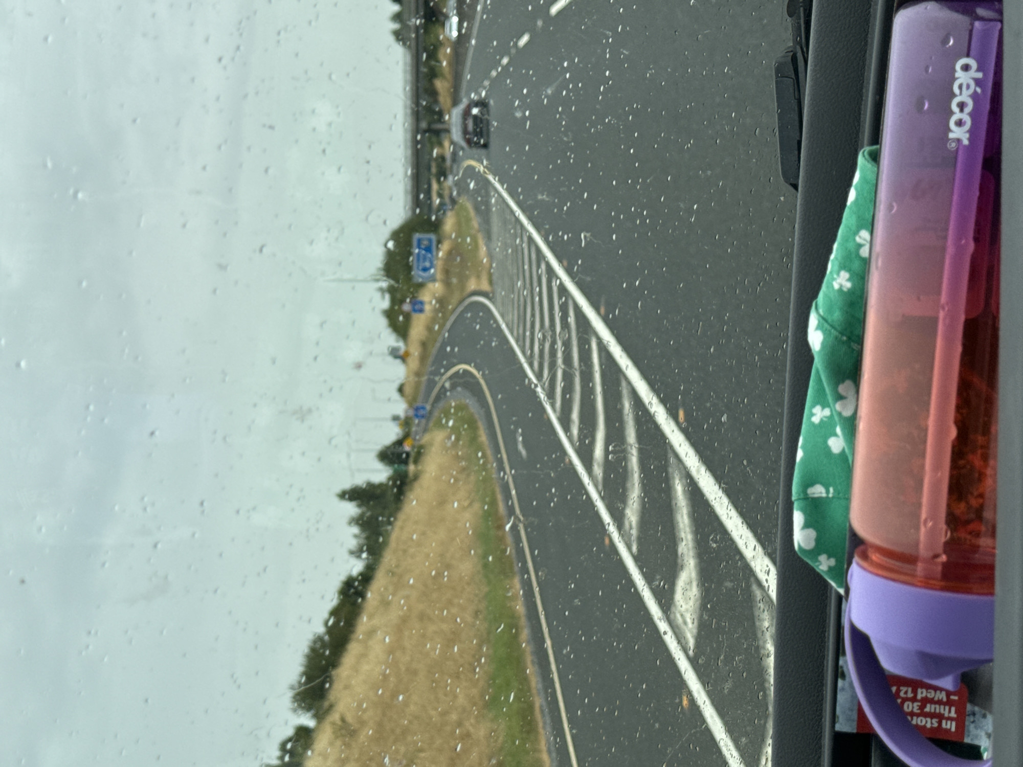 The image is a point-of-view shot from inside a vehicle, looking out through a rain-streaked windshield at a highway in Ireland. The road is wet, with visible tire tracks and white lane markings. On the right side of the road, there is a bridge overpass, and on the left, a grassy, dry hillside. Several cars are visible on the highway, some in the distance. In the foreground, on the dashboard of the vehicle, a purple and pink water bottle is visible, along with a green cloth with white shamrock patterns. The sky is overcast and grey.