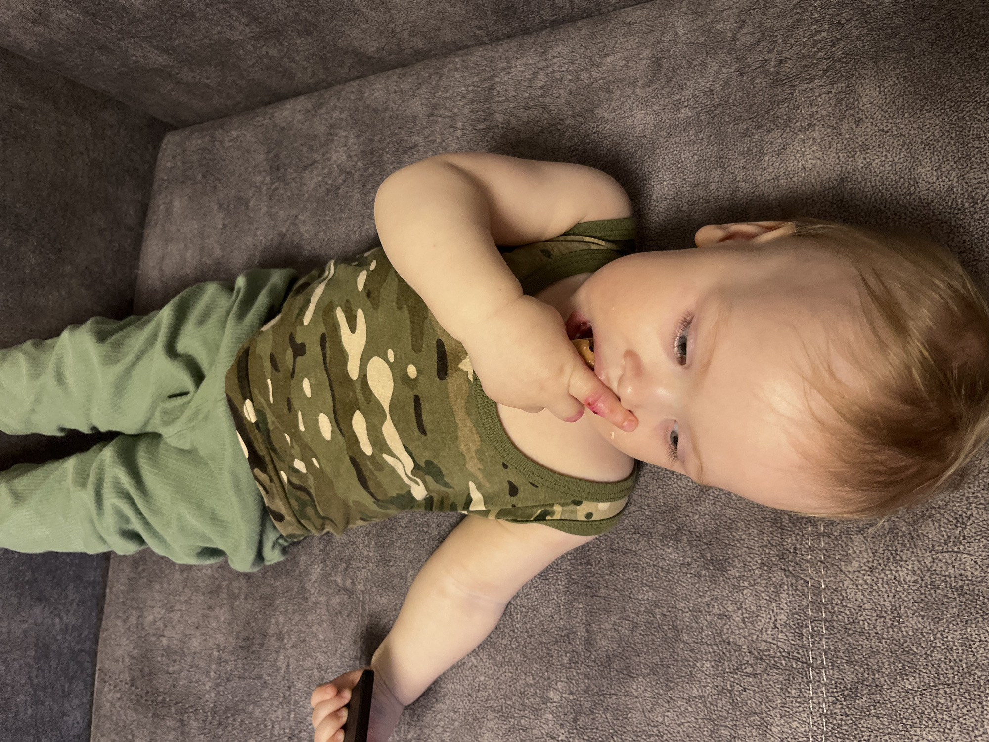 A baby is lying on a gray couch. The baby is wearing a camouflage tank top and green pants. The baby's hand is near their mouth, and there is something on their fingers. The baby is holding a black object in their other hand. The location is not discernible from the image.