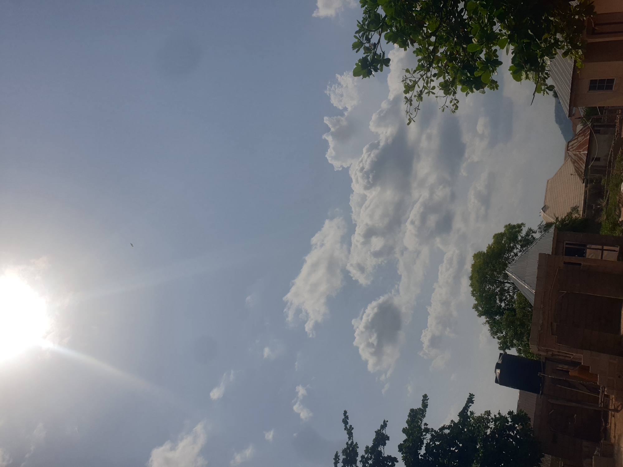 The image depicts a daytime scene in Zango-ndaji, Nigeria, featuring a low-angle view of a partly cloudy sky with the sun visible in the lower left. To the right, there are residential buildings with corrugated metal roofs and unfinished brick walls, partially obscured by trees. A black water tank is visible atop one of the buildings. The scene suggests a residential area under construction or in a state of ongoing development.