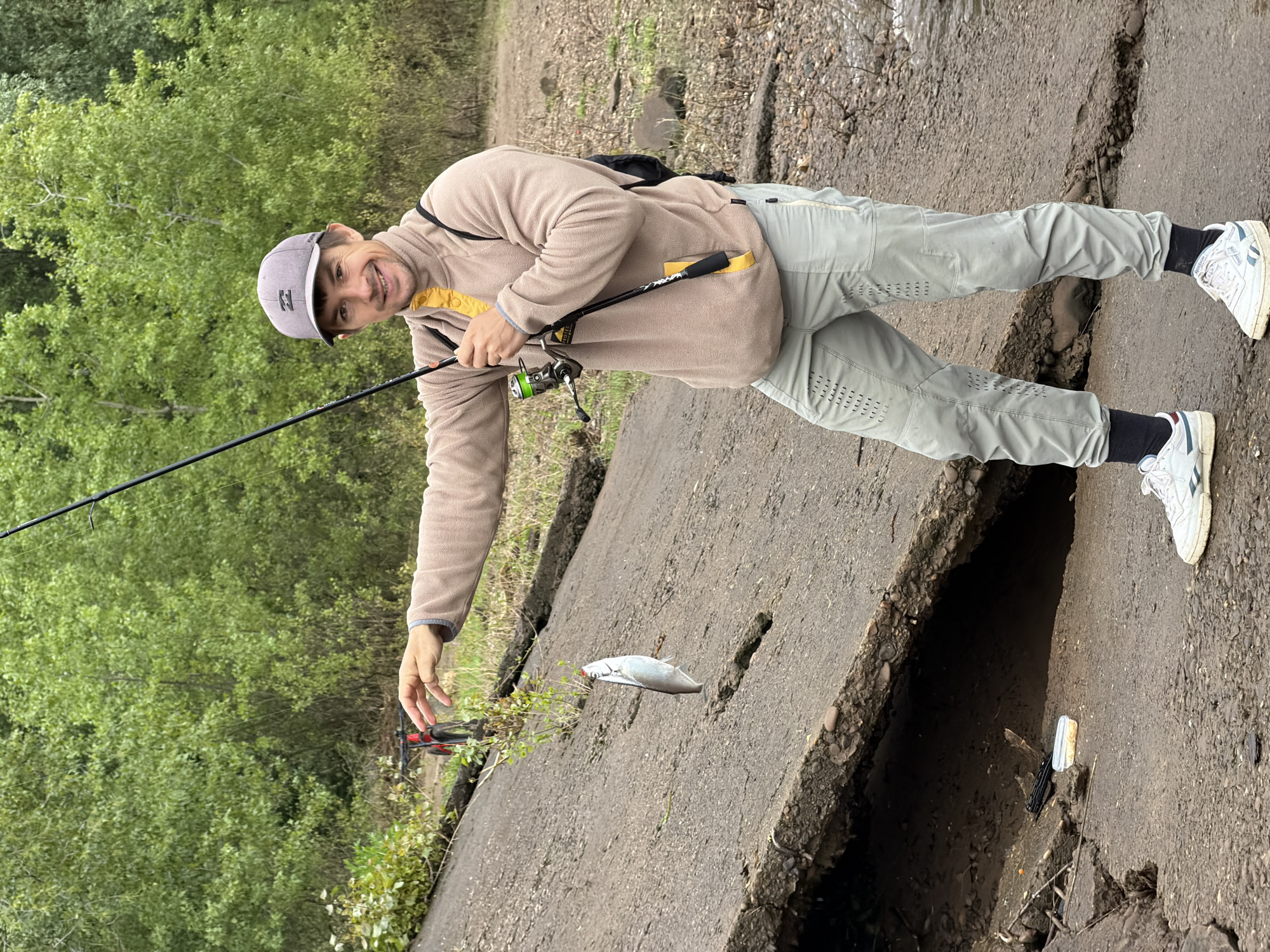 A male subject stands on an uneven ground comprising dirt and a cracked concrete structure. He is wearing a grey baseball cap with a visible 'B' logo, a beige fleece quarter-zip pullover with yellow collar and pocket accents, light grey athletic pants with perforated detailing, and white Reebok brand sneakers. The subject holds a fishing rod with a green reel in his right hand. From his extended left hand, a small, silver-scaled fish with a reddish head area is suspended by a fishing line. He is smiling and looking towards the camera. A black backpack is partially visible on his back. The background consists of dense green foliage, primarily trees. A red and black mountain bicycle is partially visible amongst the vegetation behind the subject's left arm. At the base of the damaged concrete structure, a black object resembling a handgun and a small, translucent white plastic container are present on the ground. The scene is located in an outdoor environment within the city of Noviy, Russia.